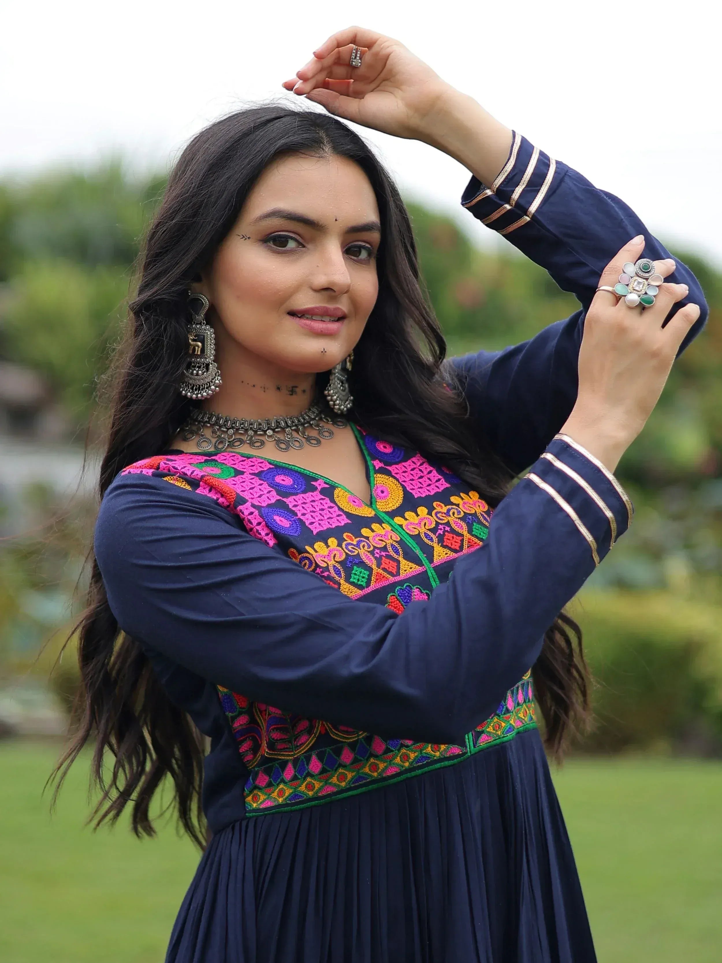 Woman in navy blue ethnic gown with vibrant embroidery, traditional jewelry, and rings, styled for Indian festive wear.