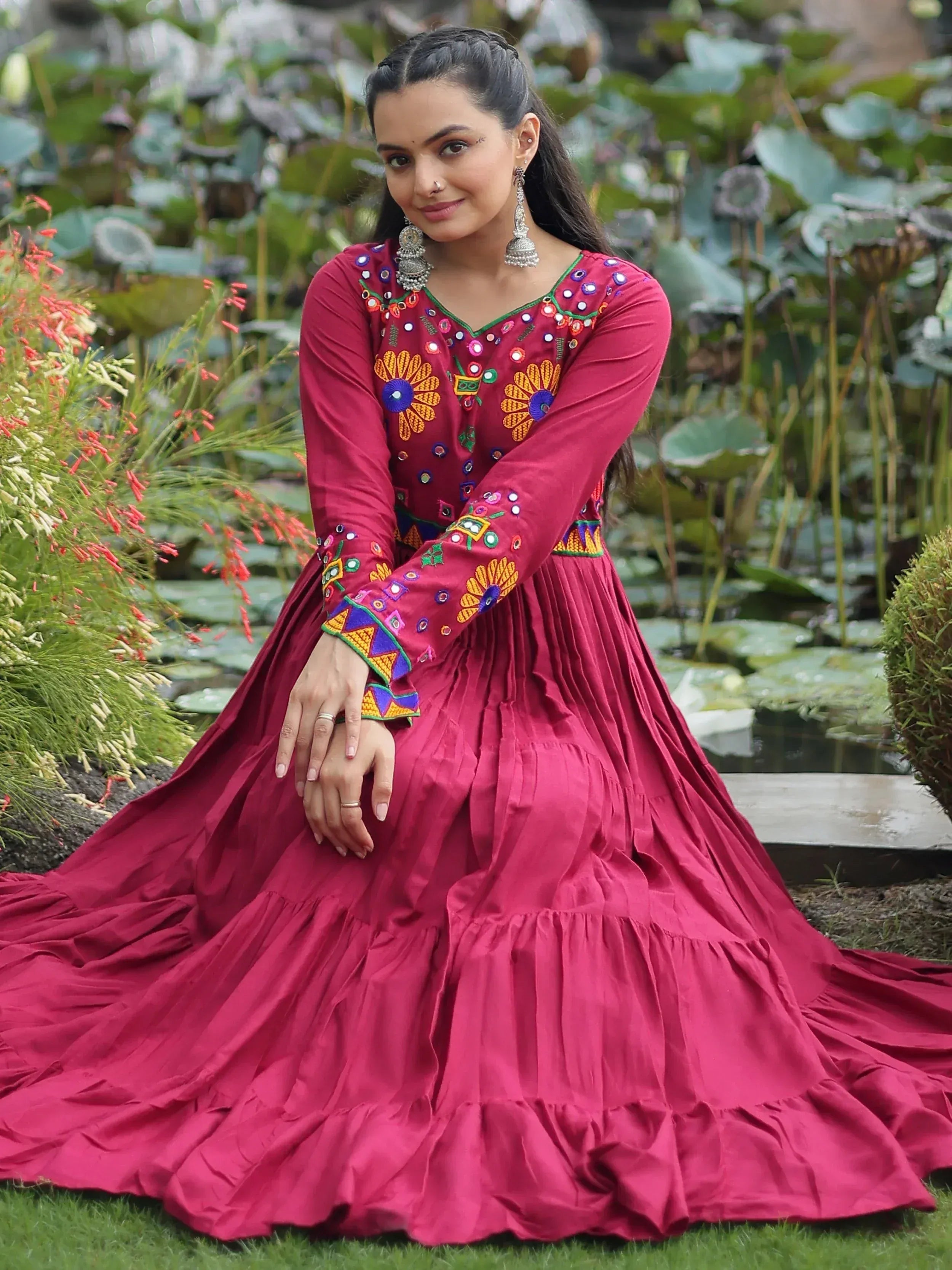 Woman in maroon ethnic gown with mirror embroidery, floral motifs, and tiered skirt, styled with jhumka earrings in garden setting