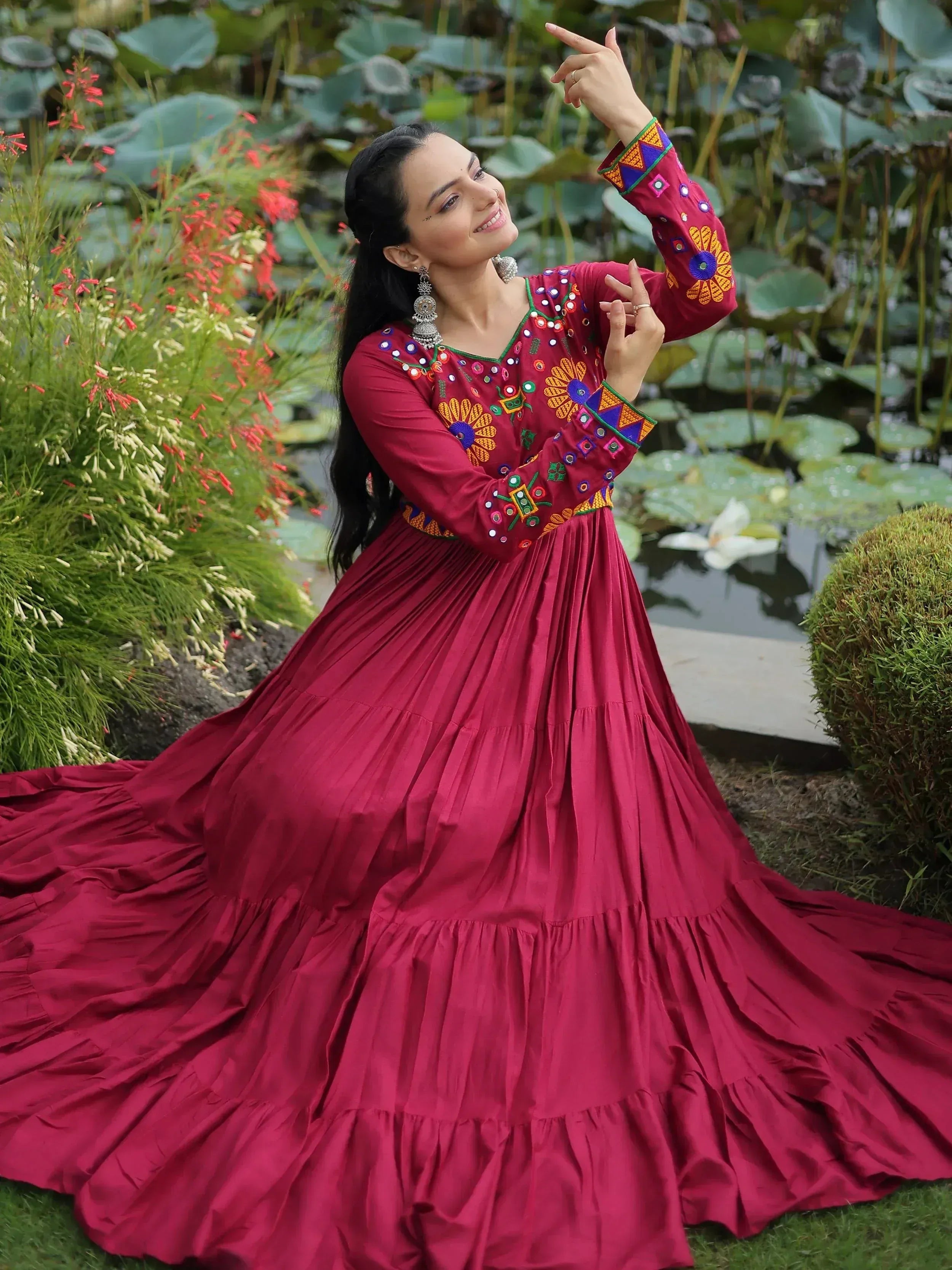 Woman in maroon embroidered ethnic gown with mirror work, posing near lotus pond