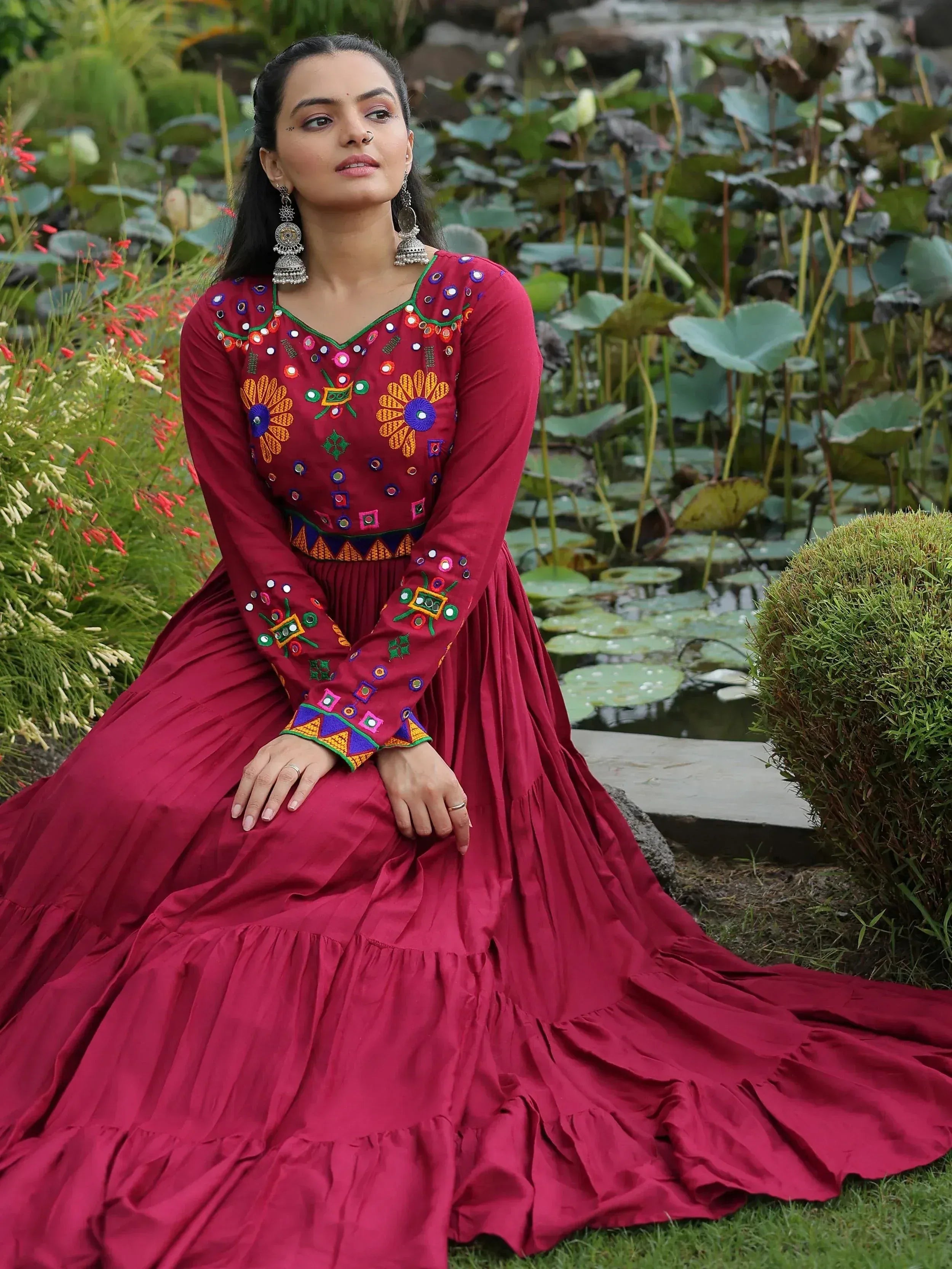 Maroon ethnic gown with mirror embroidery, floral motifs, and tiered skirt, styled with jhumka earrings in garden setting