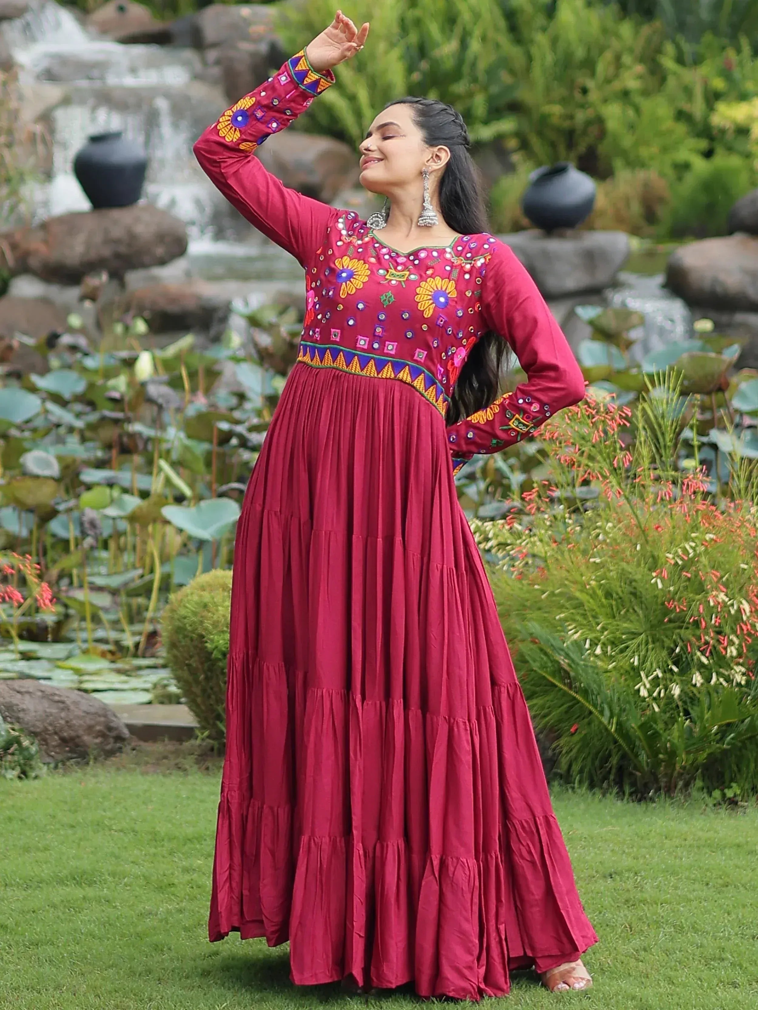 Woman in maroon embroidered ethnic gown with floral motifs, standing in garden near waterfall