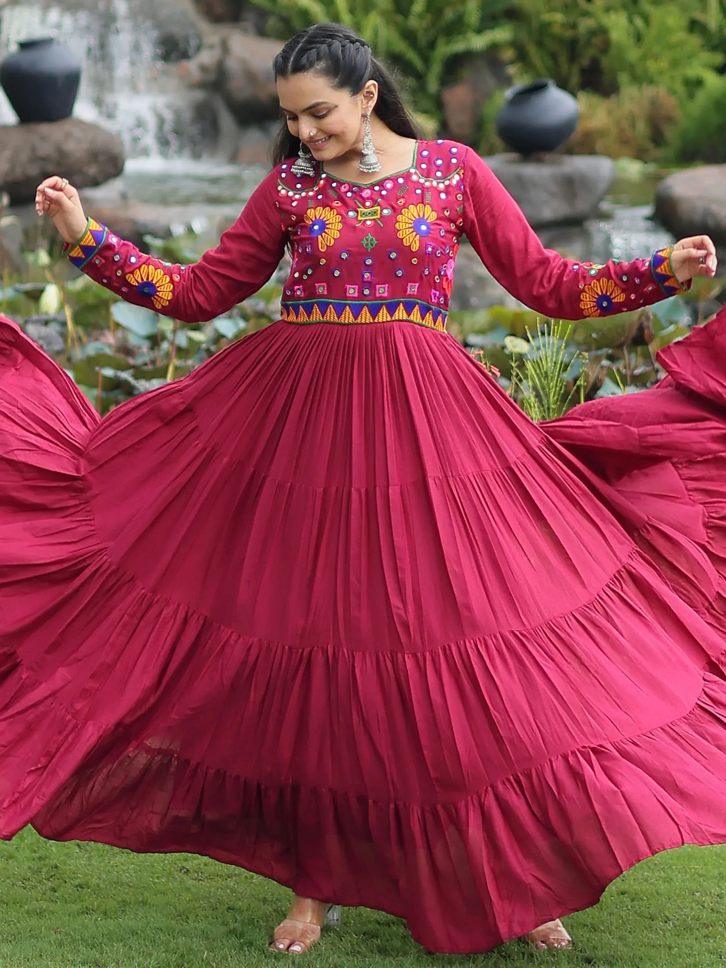 Woman twirling in maroon ethnic gown with mirror work, floral embroidery, and pleated skirt, perfect for festive occasions.