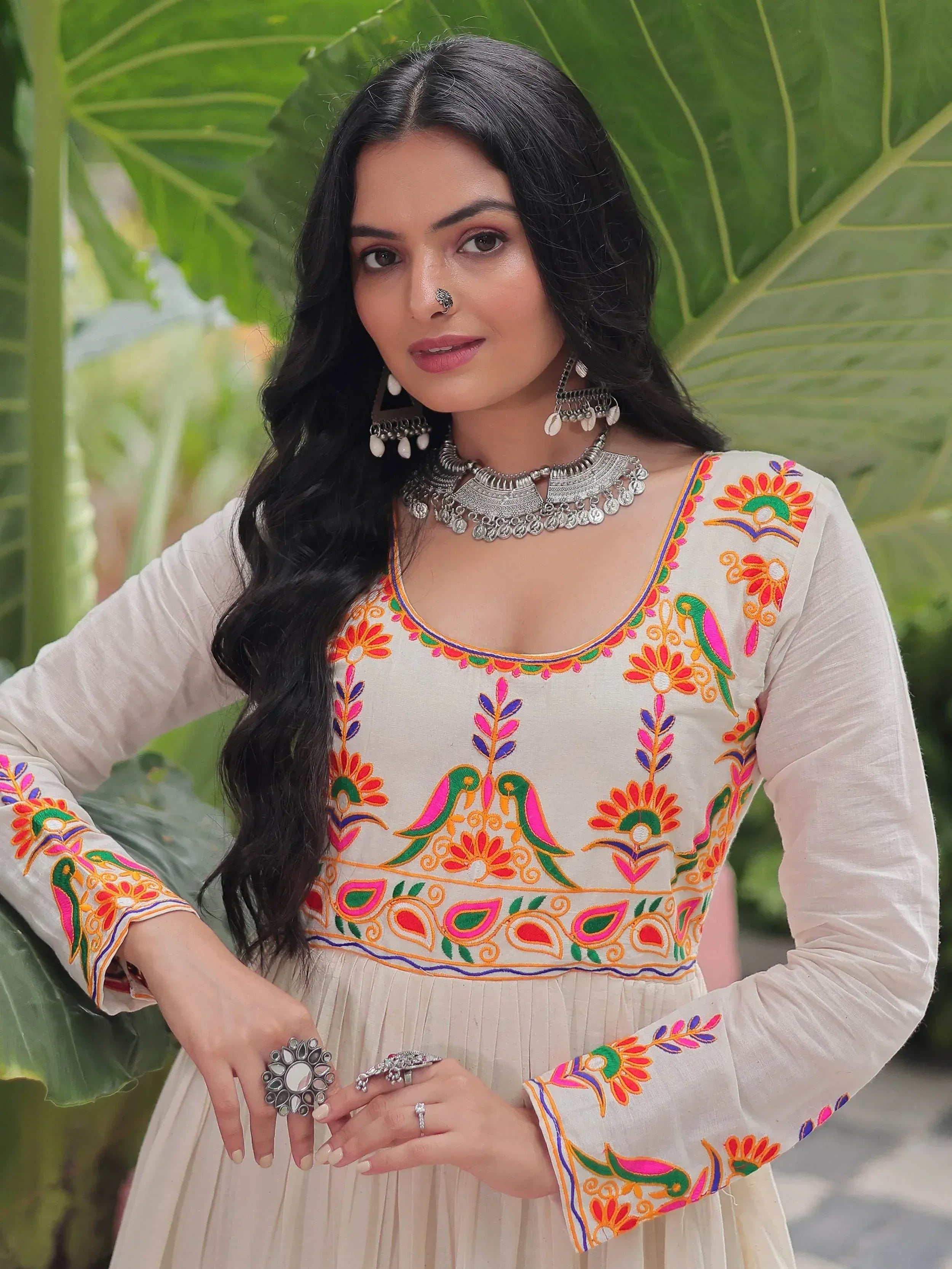 Woman in white embroidered ethnic gown with colorful floral and bird motifs, silver jewelry, green leaf backdrop