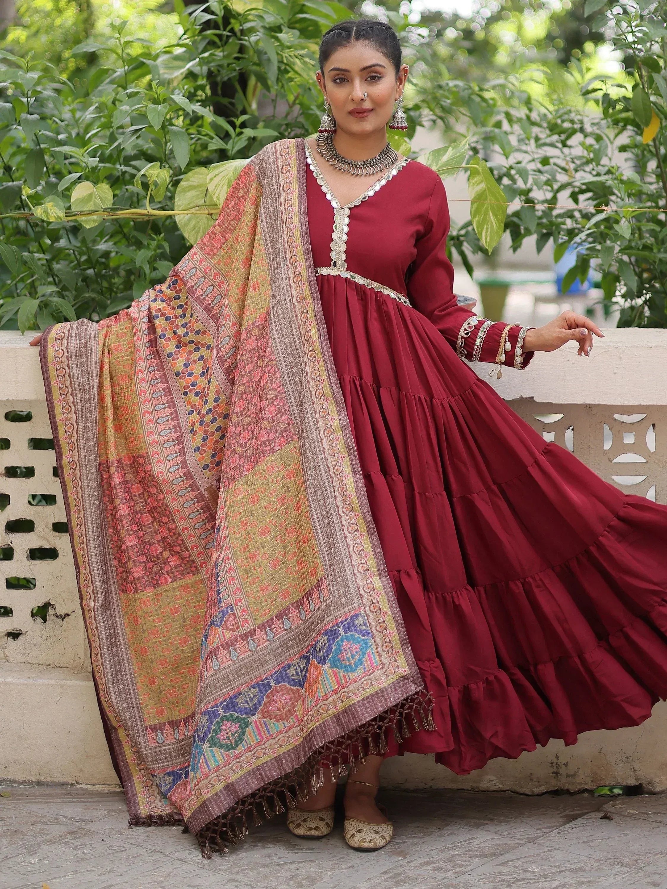 Maroon tiered ethnic gown with mirror work and printed dupatta, styled with traditional jewelry for festive occasions
