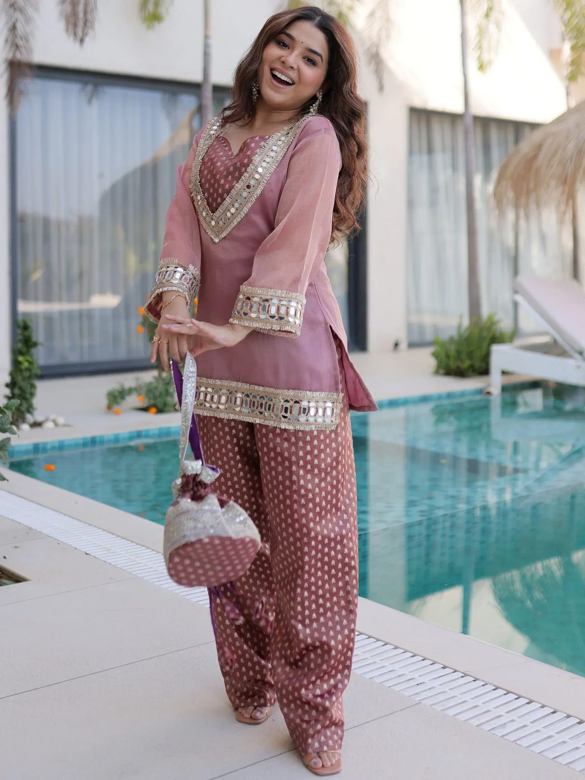 Woman in pink ethnic salwar kameez with mirror work, standing by pool, holding matching potli bag