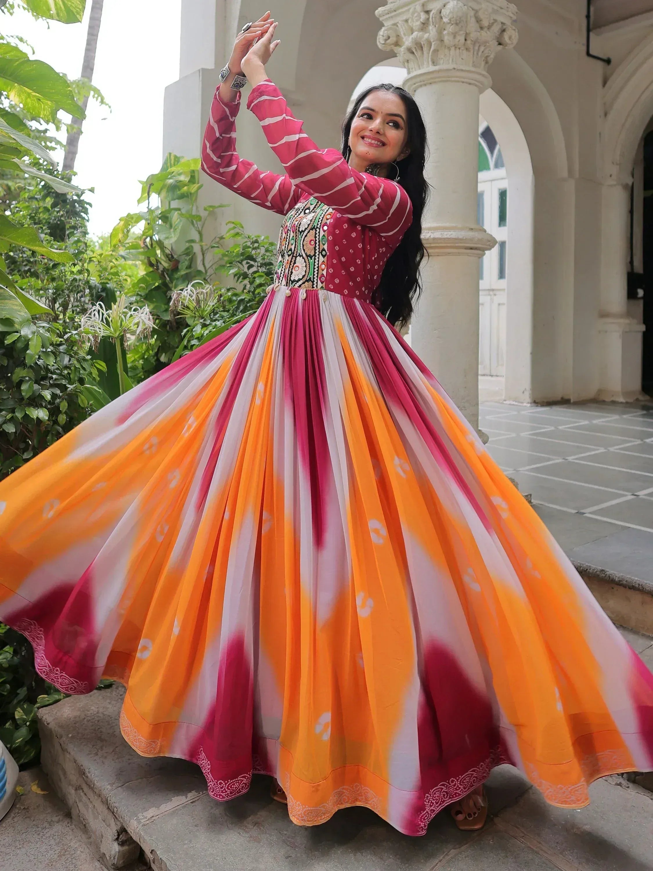 Woman in vibrant pink and orange ethnic gown with embroidered top, twirling outdoors near white pillars.