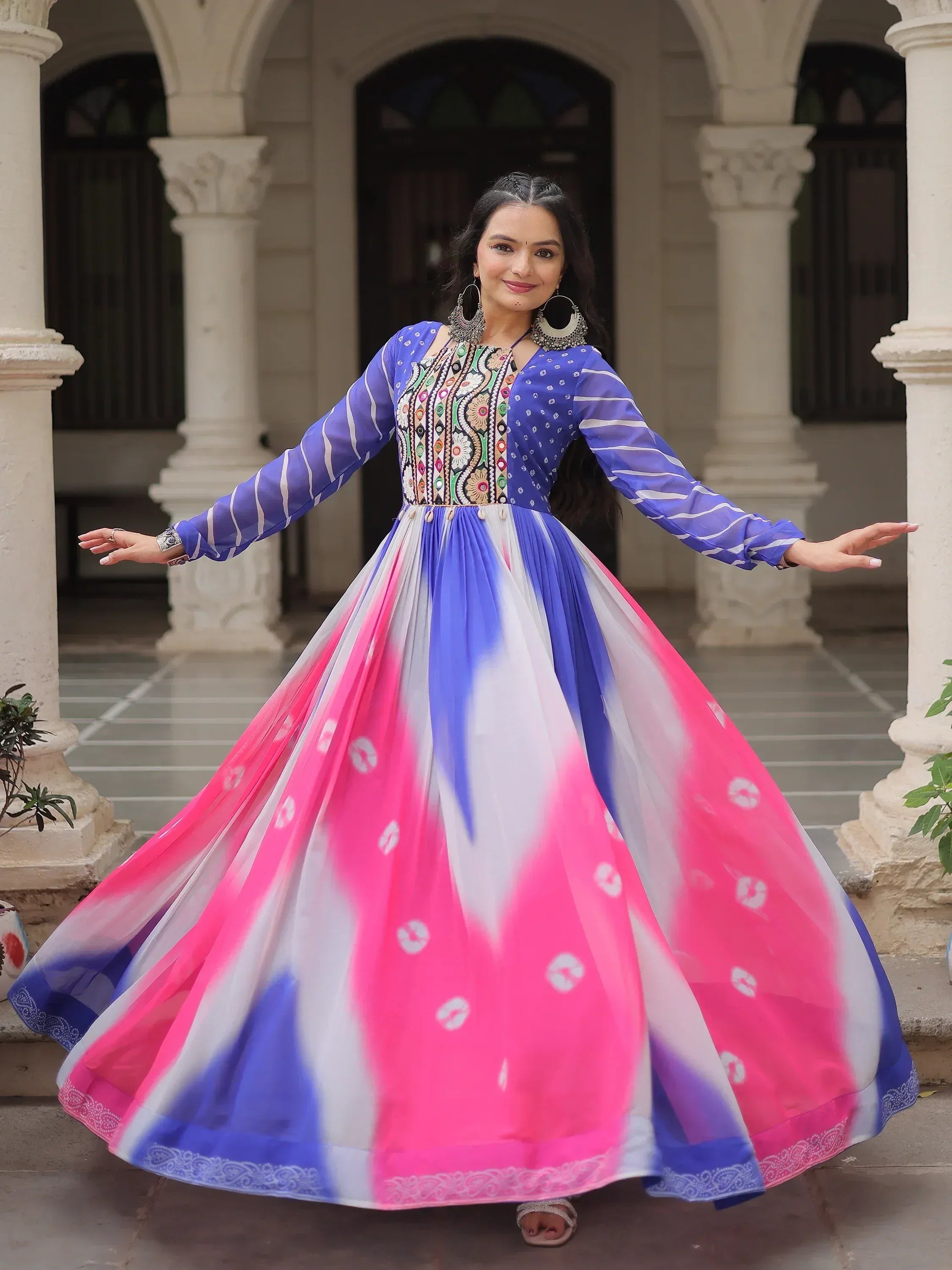 Woman in vibrant blue and pink ethnic gown with embroidered bodice, twirling in courtyard
