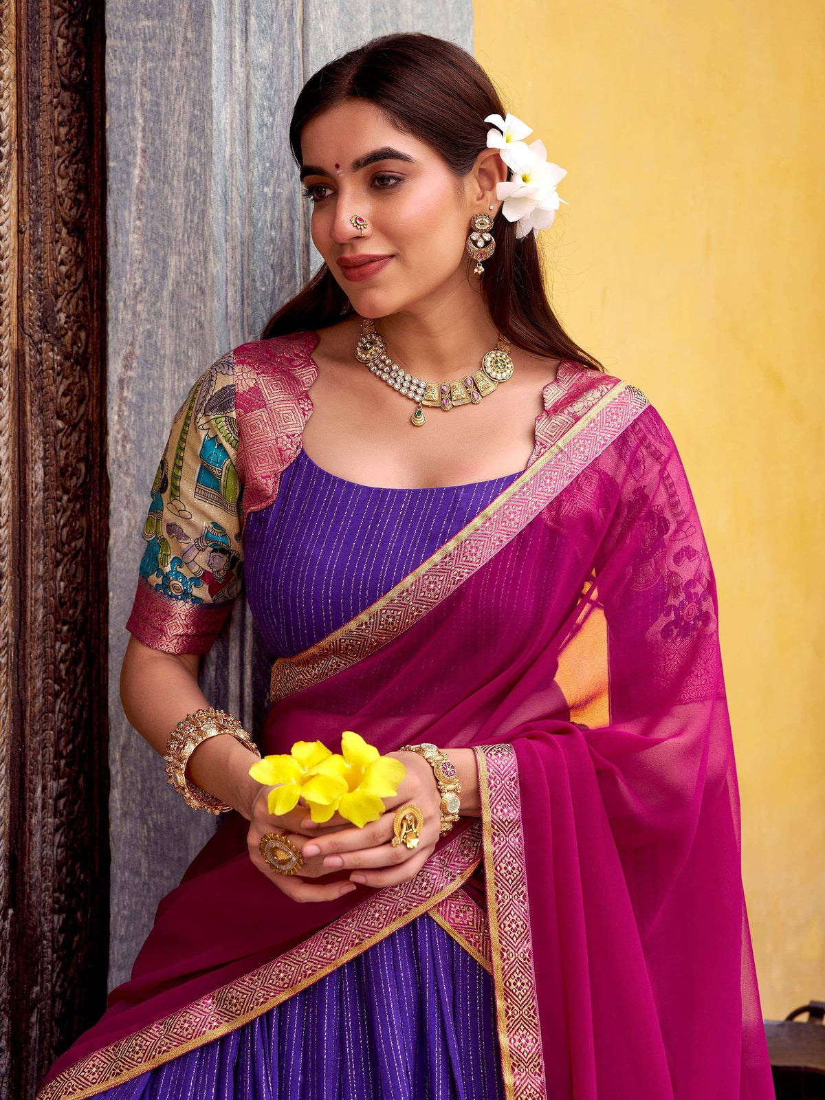 Woman in purple and magenta saree with gold border, traditional jewelry, holding yellow flowers