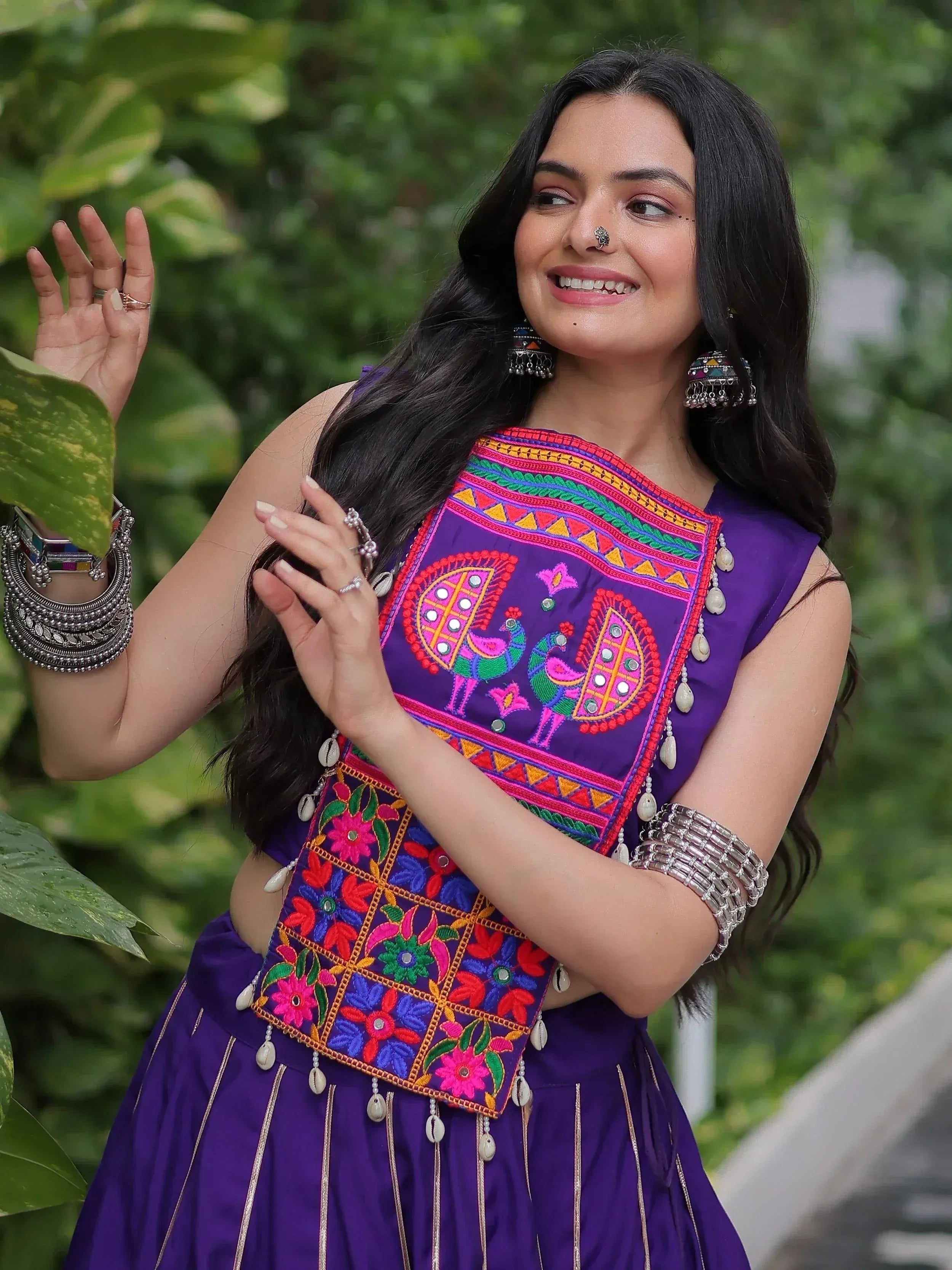 Woman in vibrant purple ethnic wear with peacock embroidery, floral patterns, and mirror work, accessorized with silver jewelry.