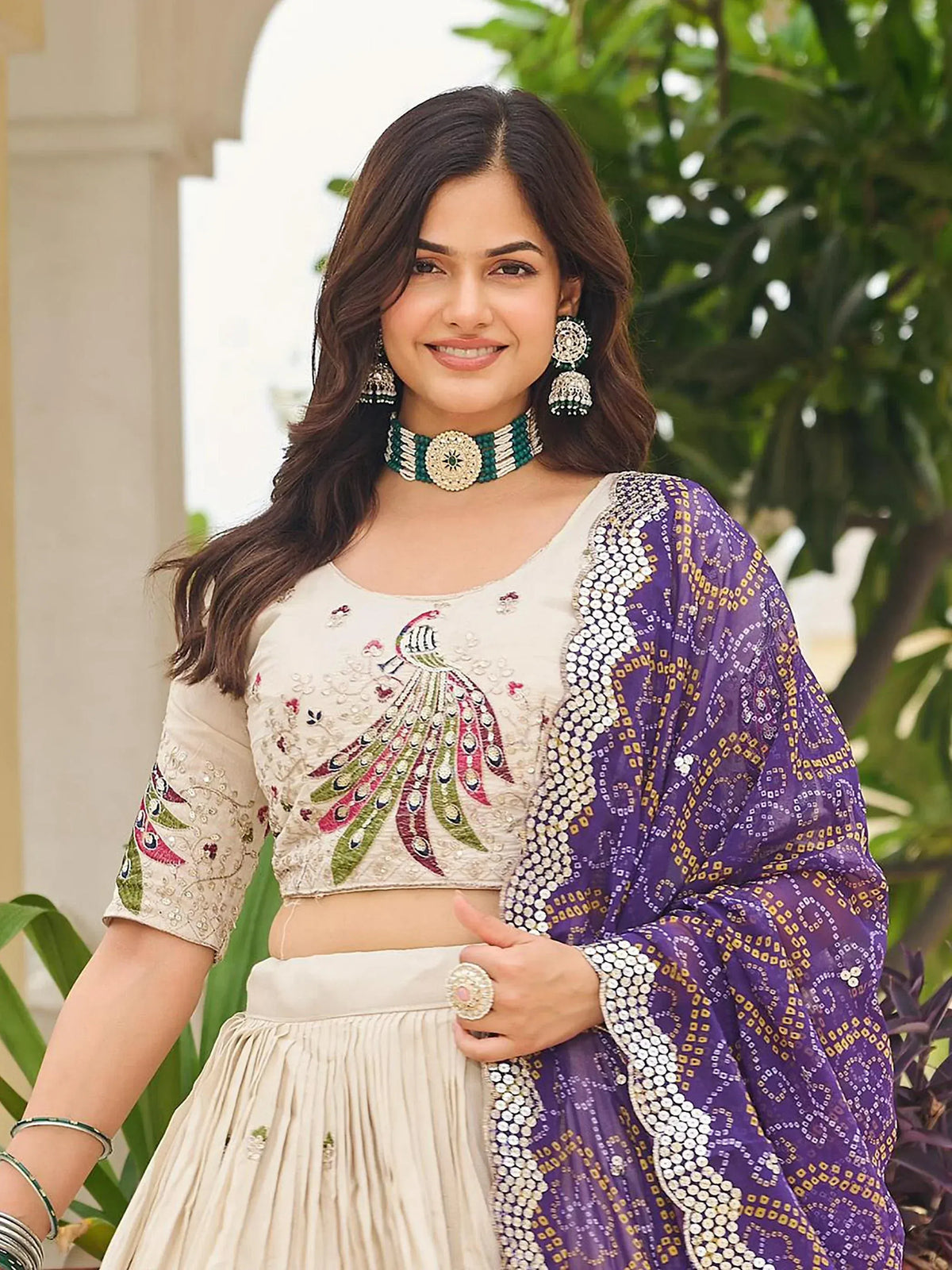 Woman in embroidered beige lehenga choli with peacock design, paired with purple sequin dupatta and traditional jewelry
