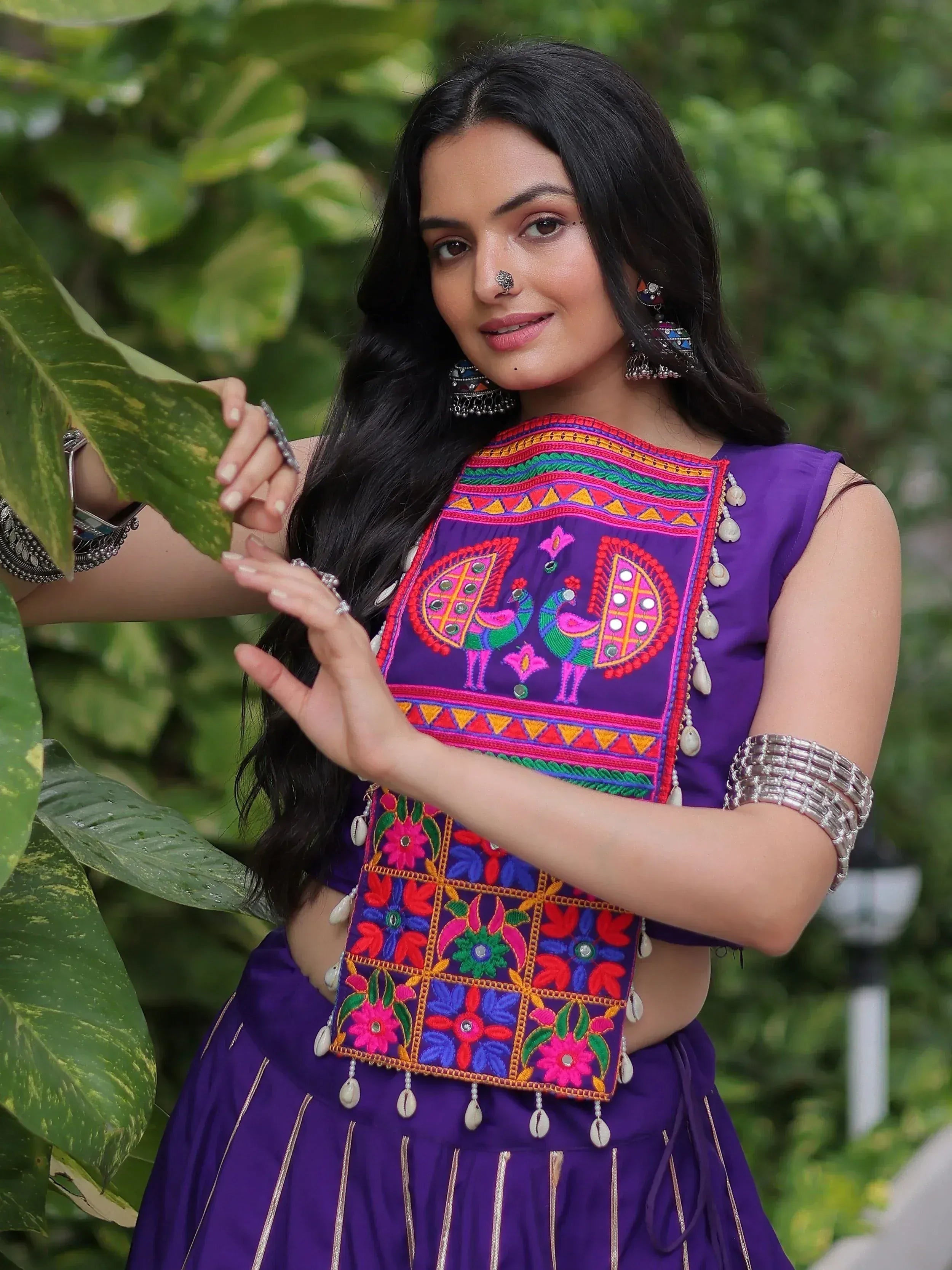 Woman in purple ethnic wear with peacock embroidery, floral patterns, and shell embellishments, styled with traditional jewelry.