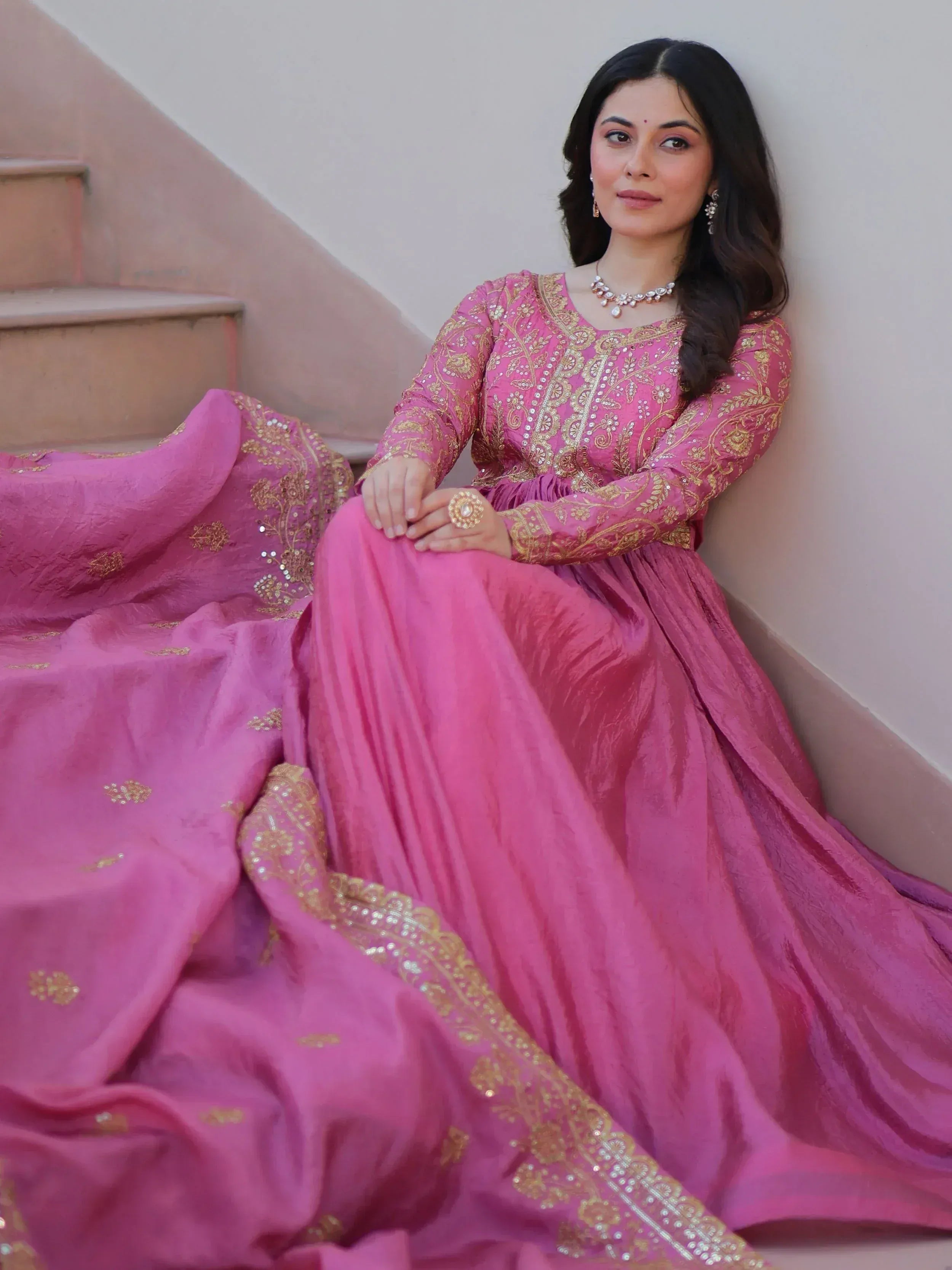 Woman in pink embroidered ethnic gown with gold detailing, sitting on stairs