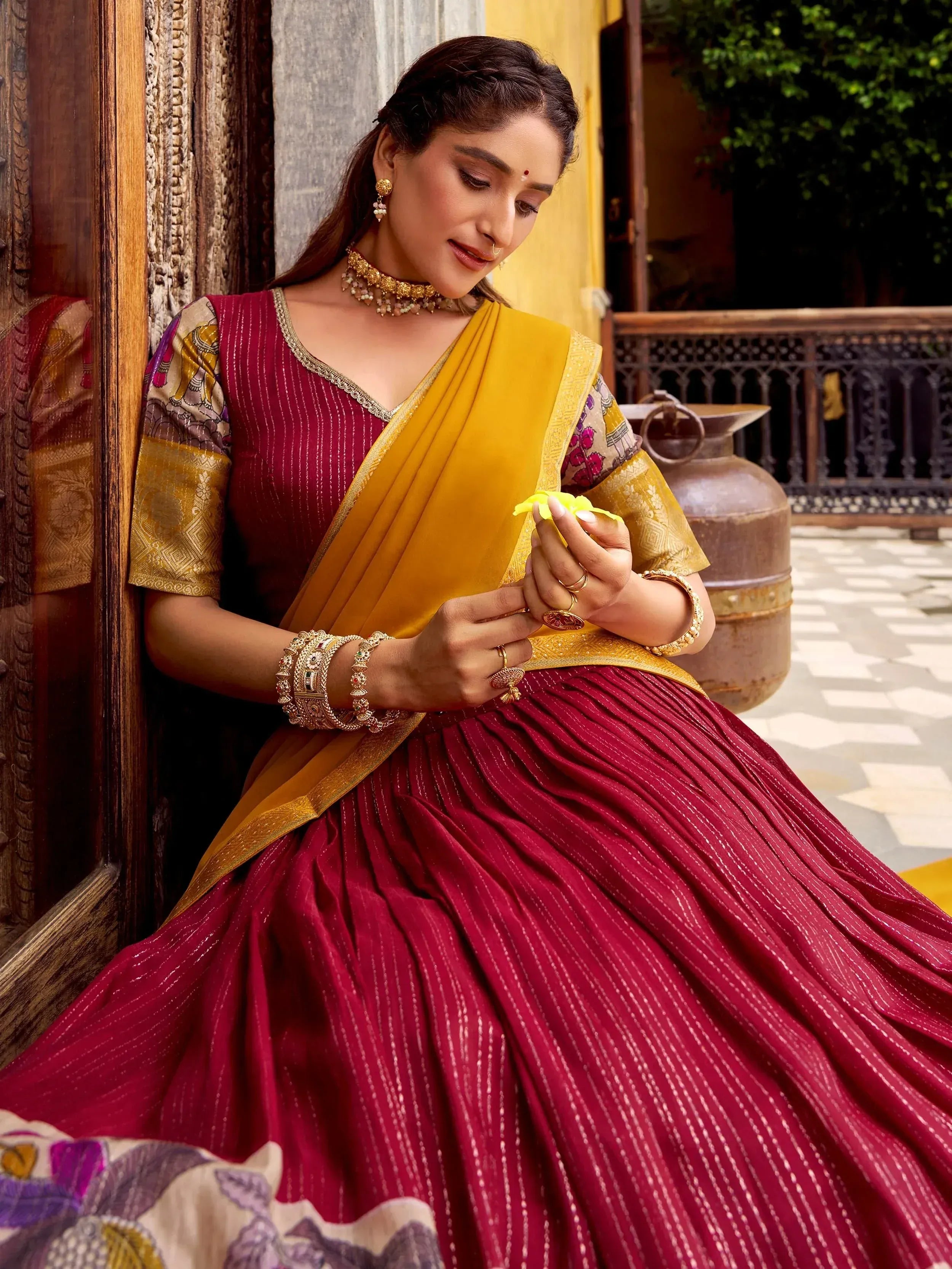 Woman in maroon and yellow designer lehenga choli with gold jewelry, holding flower