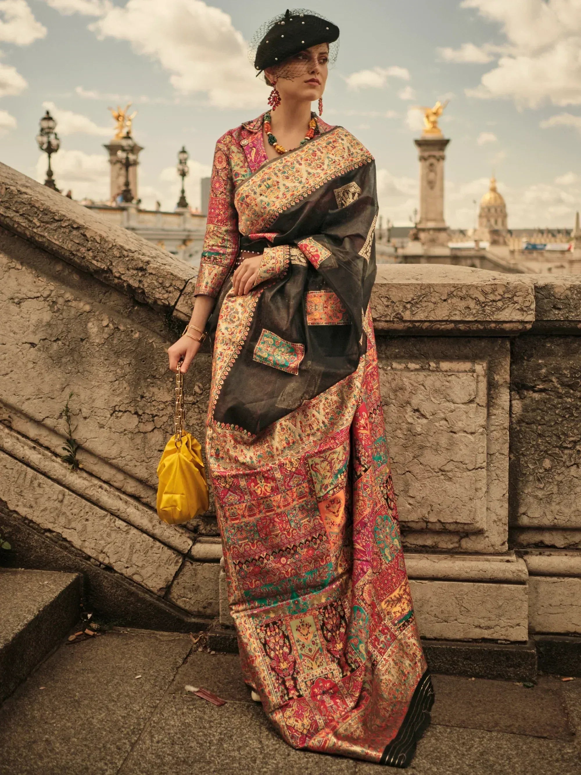 Woman in colorful printed saree with black blouse, yellow bag, and beret, standing on stone steps outdoors.