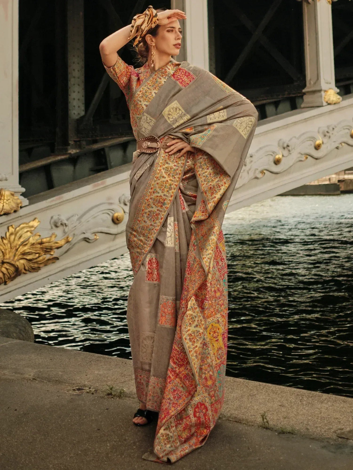 Elegant woman in designer saree with intricate patterns, standing by a bridge near water.