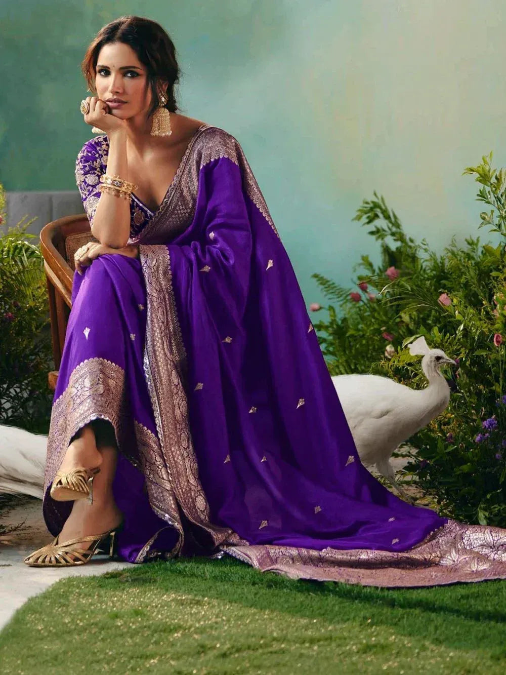 Elegant purple designer saree with gold embroidery, paired with traditional jewelry and heels, set against a garden backdrop.