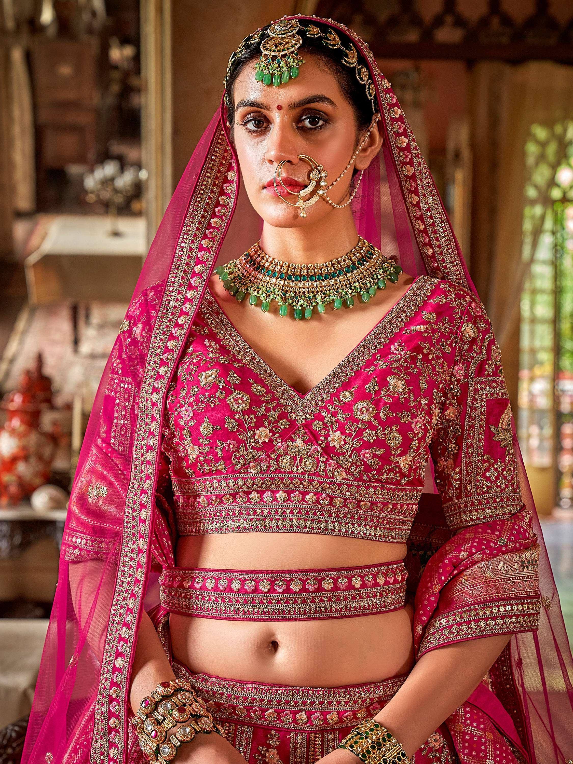 Pink embroidered lehenga choli with gold detailing, green jewelry, and traditional Indian bridal accessories
