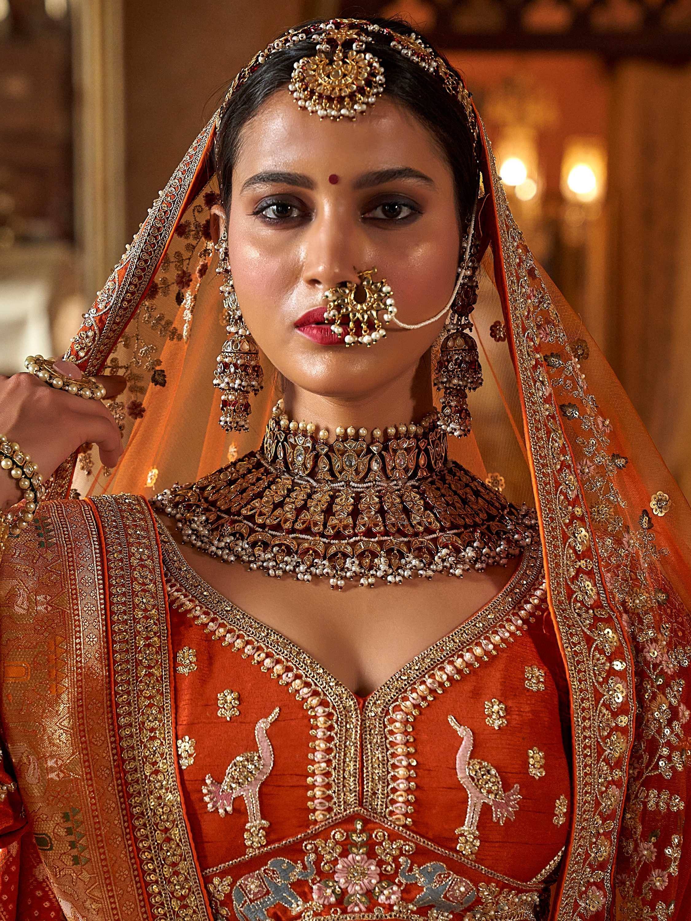 Indian bride in ornate orange lehenga choli with heavy gold jewelry and floral embroidery
