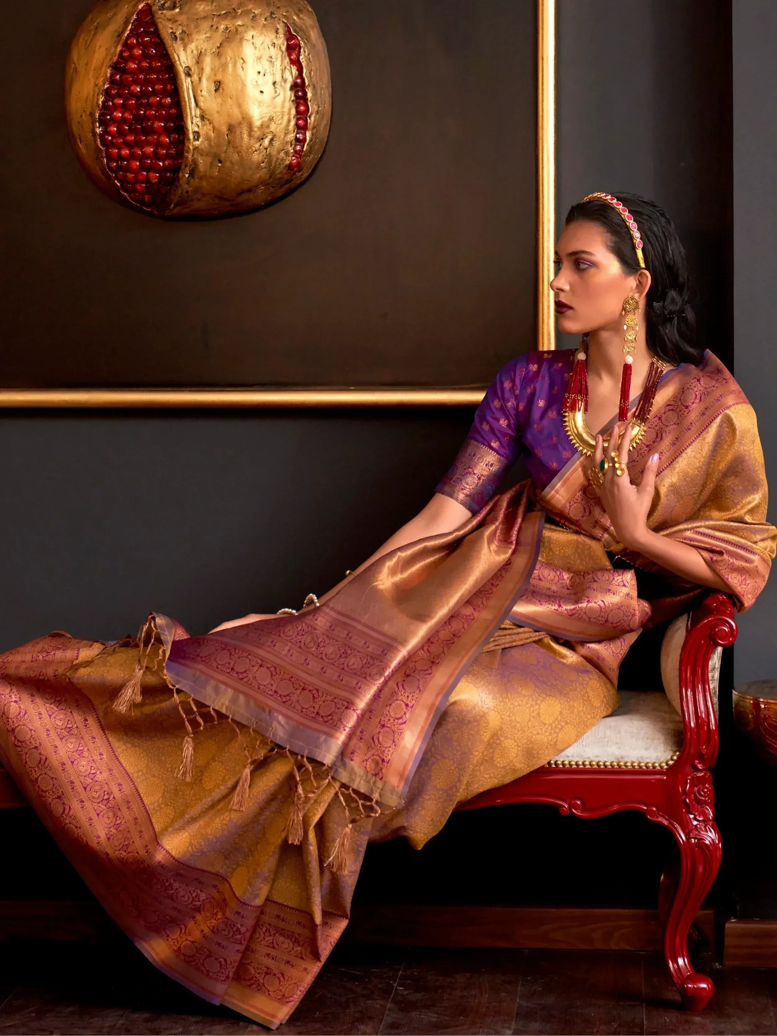 Elegant woman in purple and gold designer saree with intricate patterns, traditional jewelry, seated on red chair