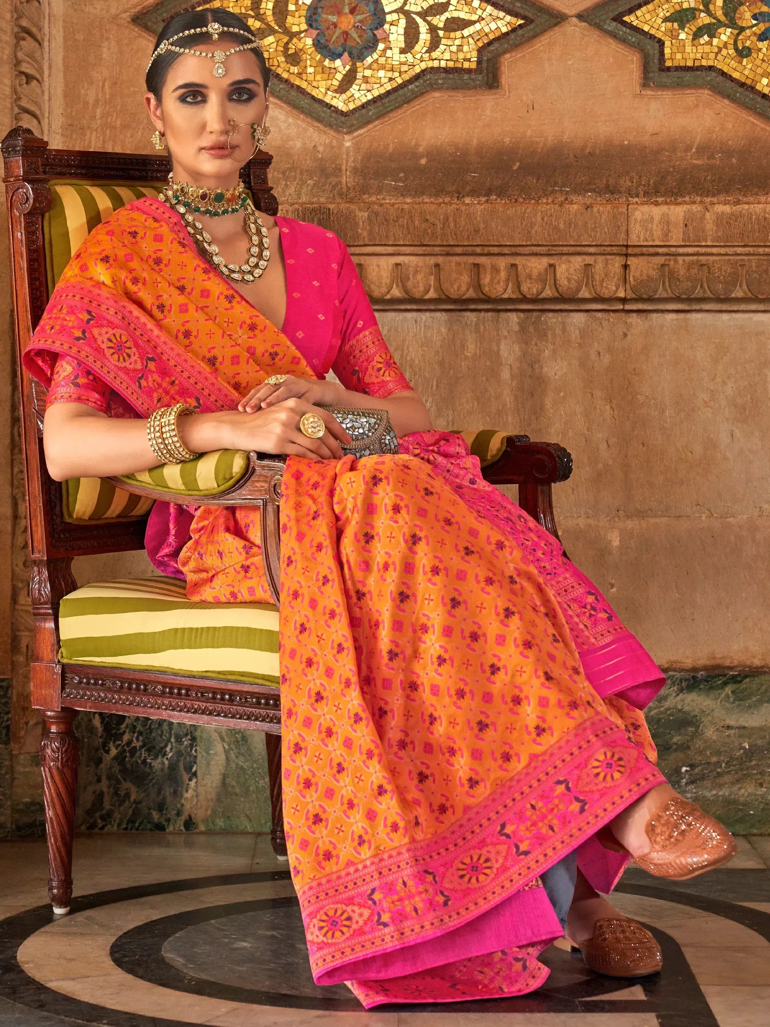 Woman in vibrant orange and pink printed saree with traditional jewelry, seated on ornate chair
