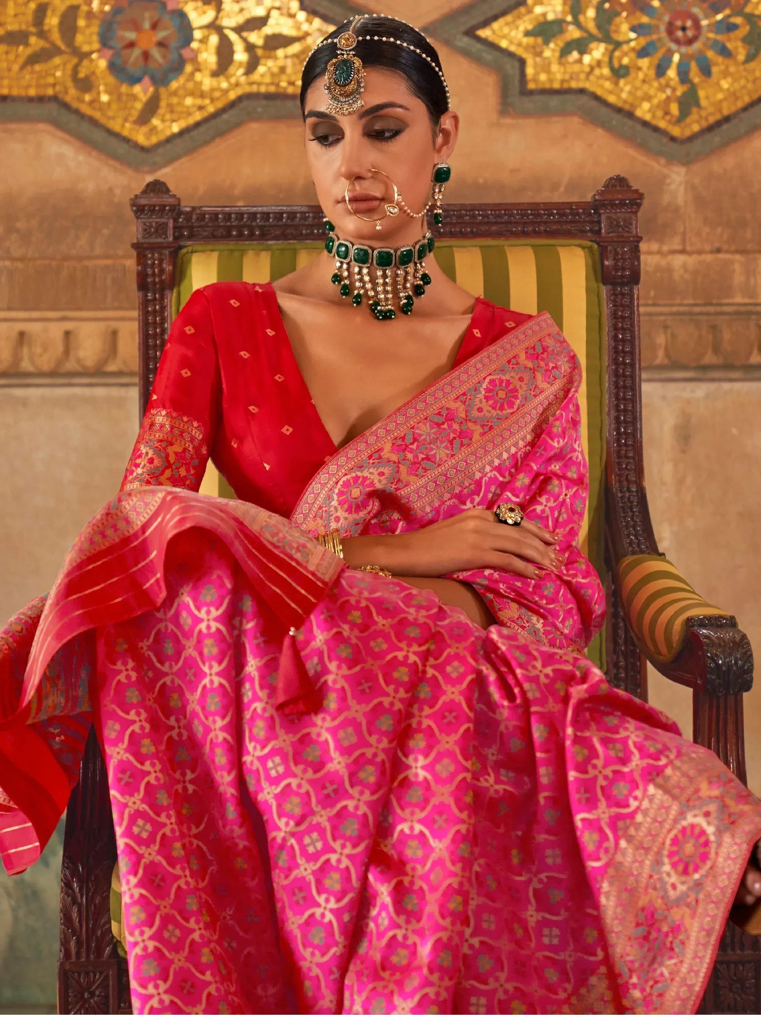 Woman in vibrant pink and red designer lehenga choli with intricate gold embroidery, traditional Indian jewelry, seated on ornate chair