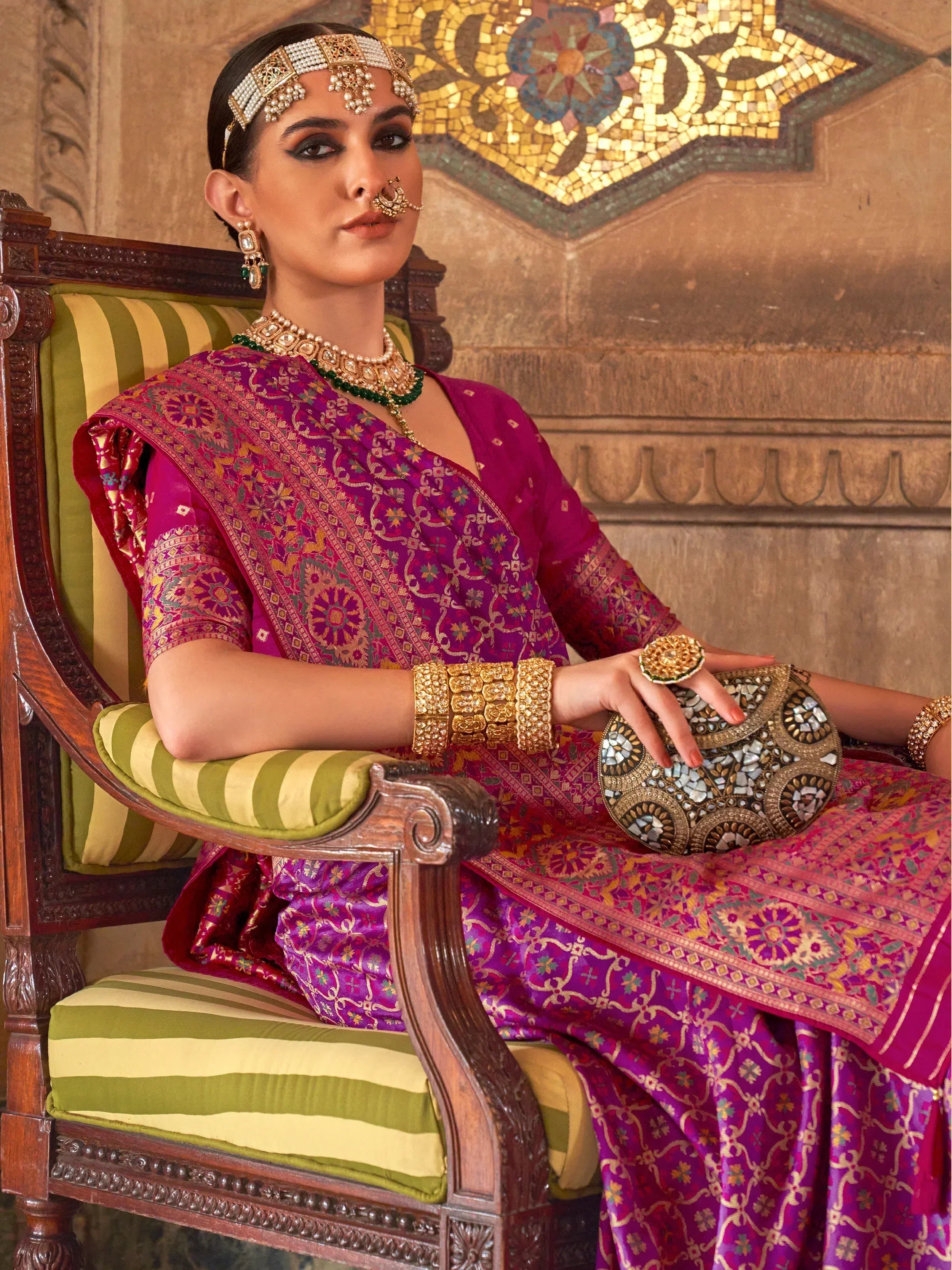 Woman in purple designer saree with gold embroidery, traditional Indian jewelry, and mirror-work clutch, seated on carved chair.