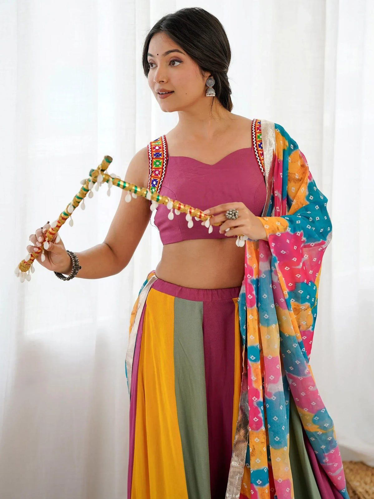 Woman in colorful Indian ethnic wear with embroidered top, striped skirt, and tie-dye dupatta holding decorative stick