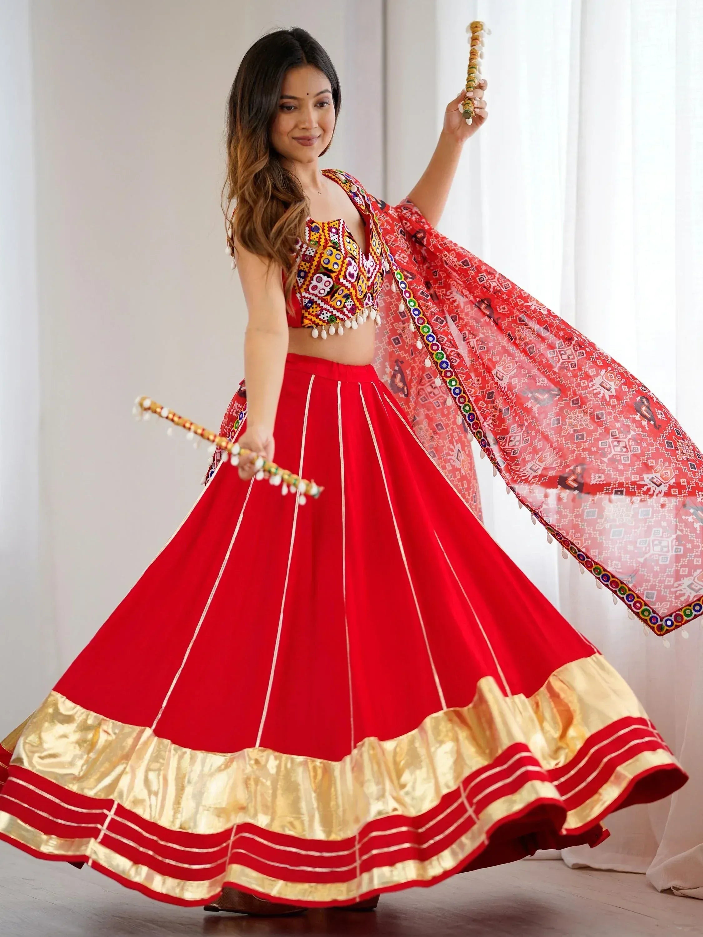 Woman in vibrant red lehenga choli with mirror work, dancing with dandiya sticks, perfect for Indian festivals.