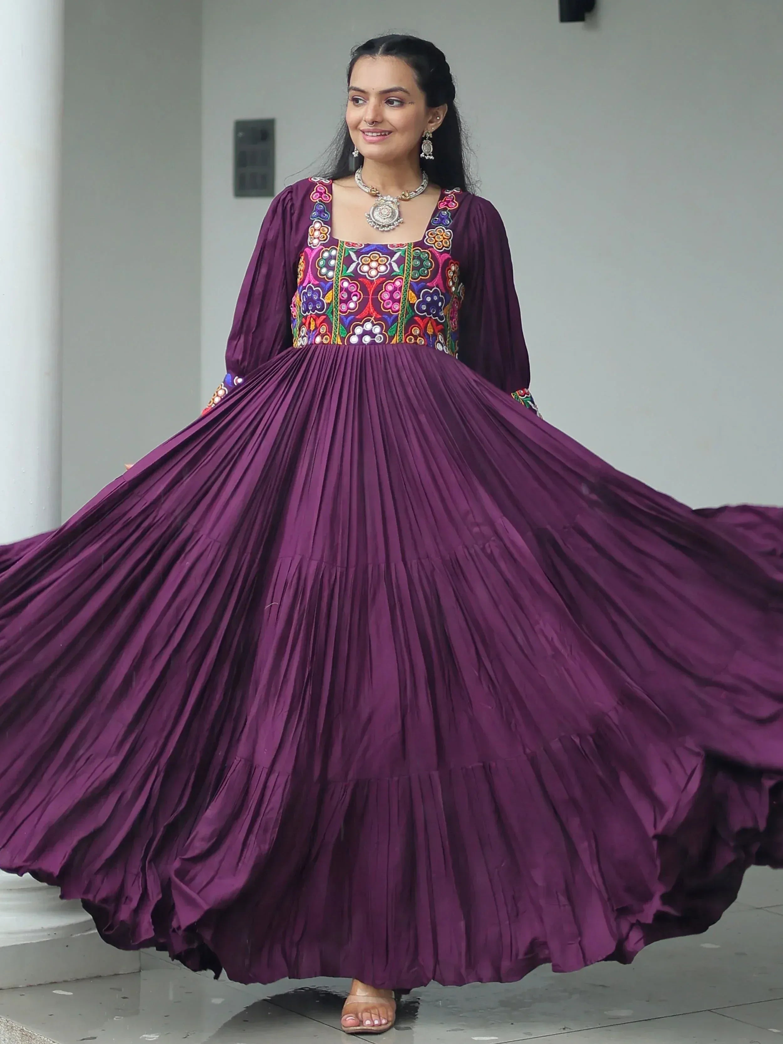 Woman in purple ethnic gown with colorful floral embroidery and pleated skirt, styled with traditional jewelry