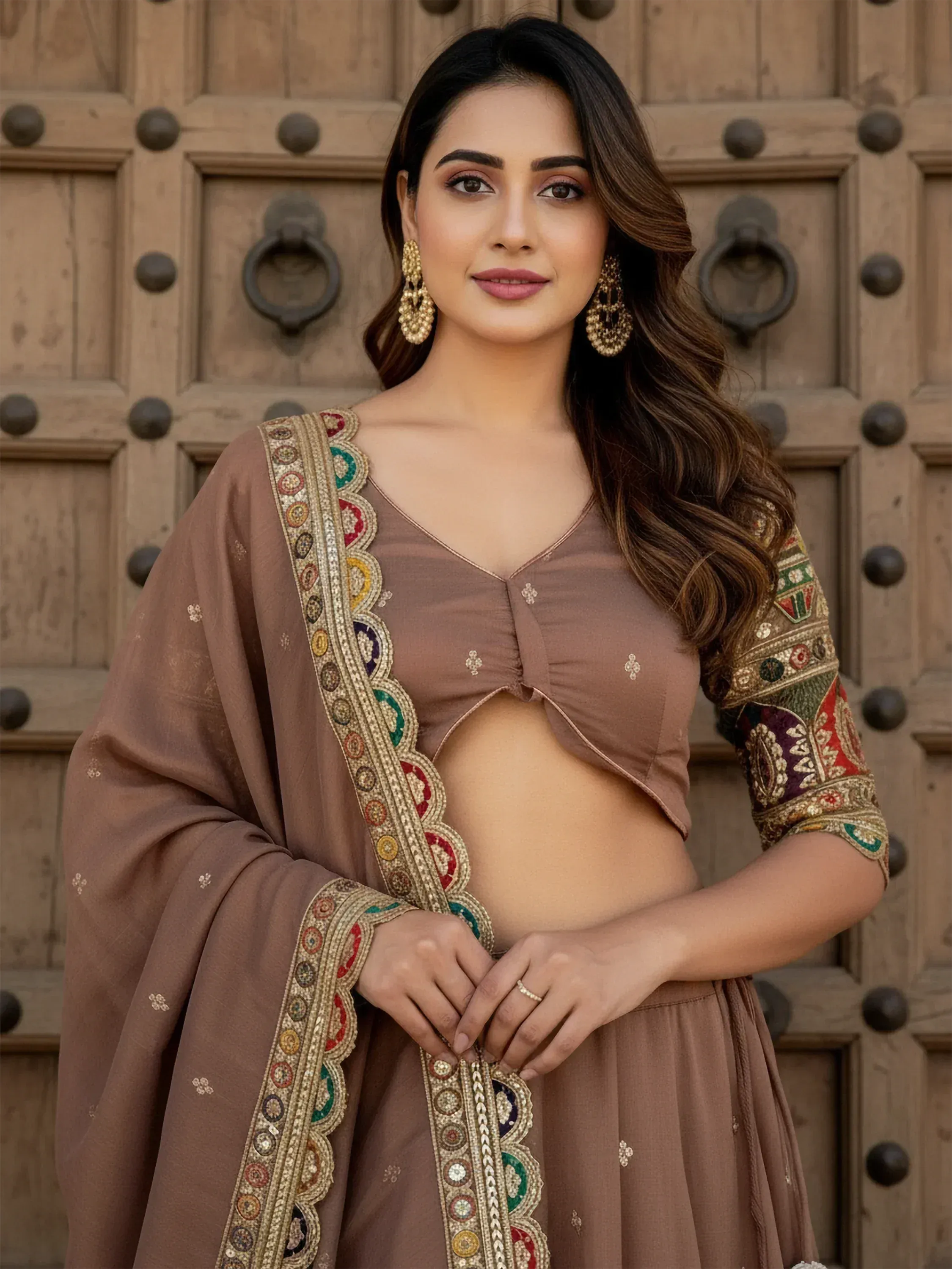 Woman in elegant brown ethnic lehenga choli with colorful embroidery, standing before a rustic wooden door.