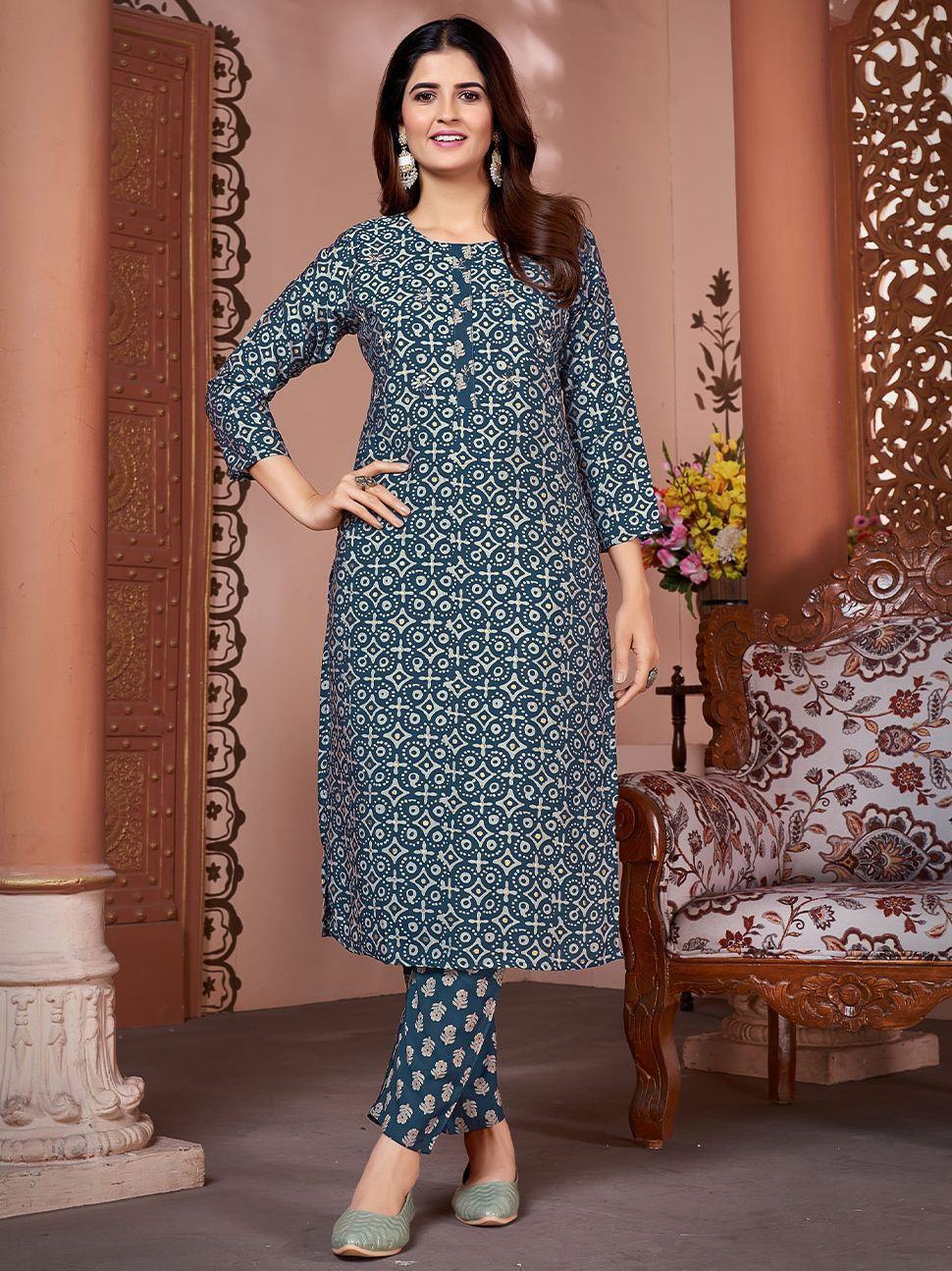 Woman in blue printed kurta set with ethnic patterns, standing indoors next to a floral chair
