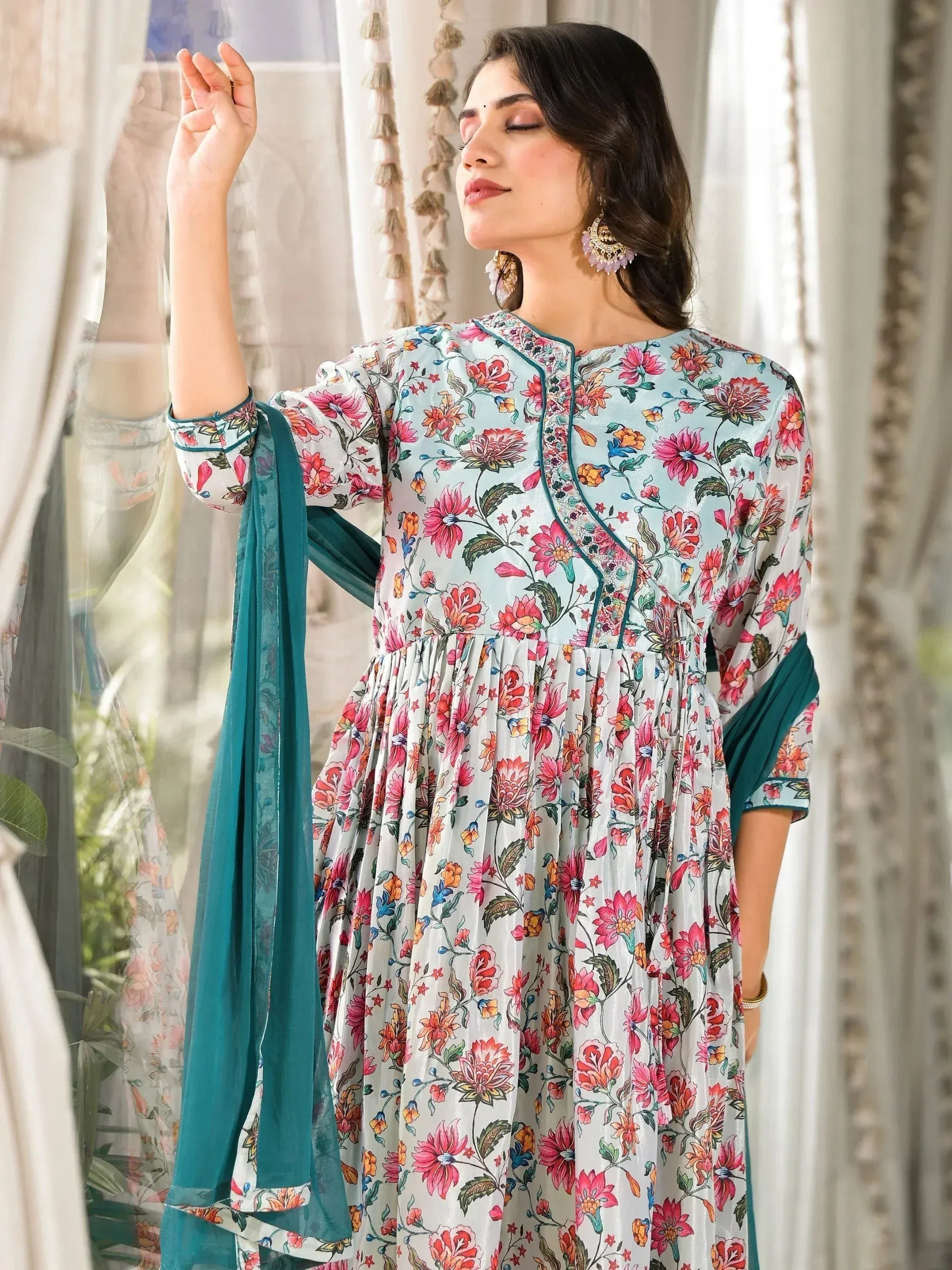 Woman wearing a floral print ethnic gown with teal dupatta near window curtains.
