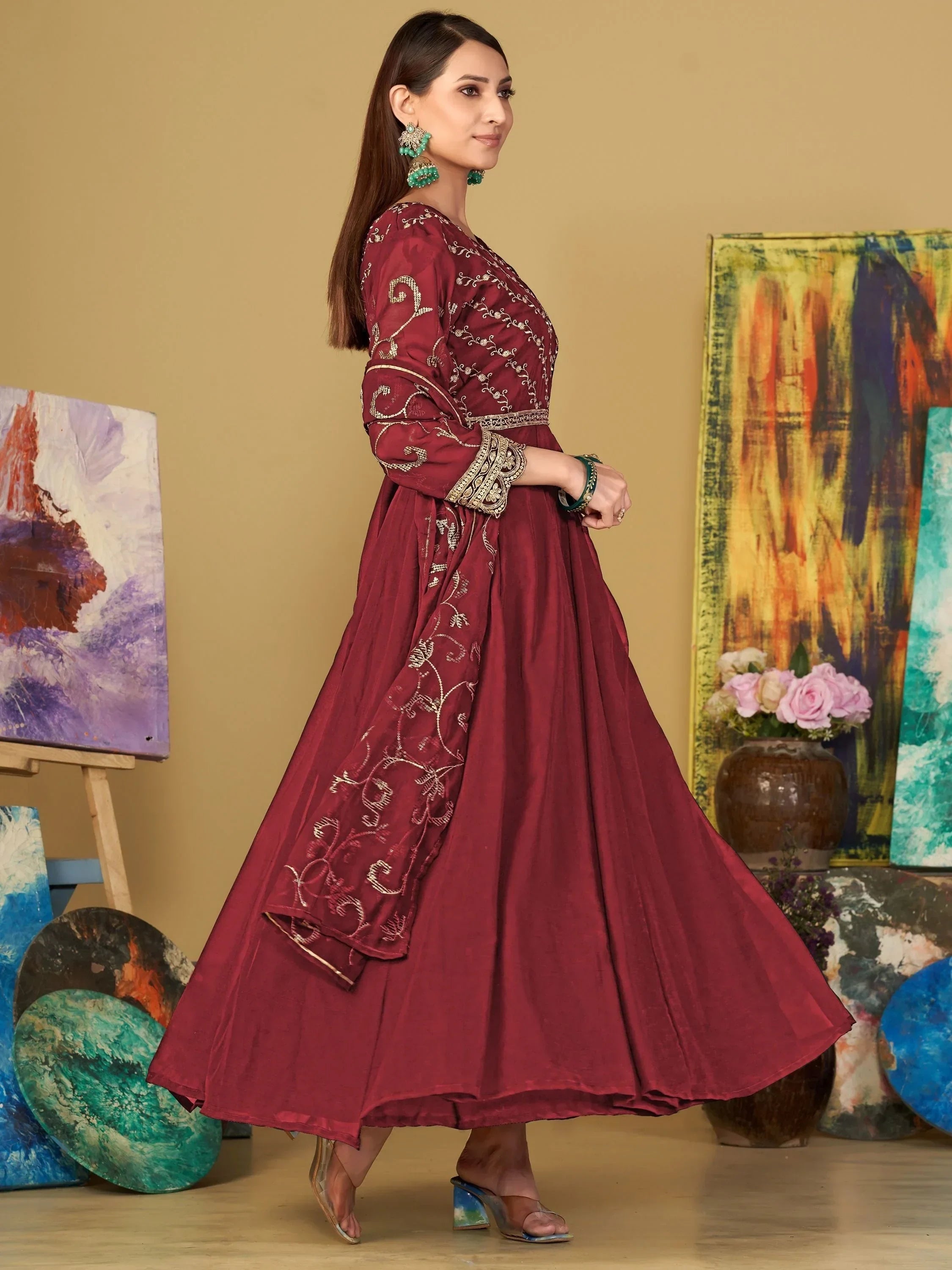 Woman in a maroon embroidered ethnic gown with sheer sleeves, posing indoors near colorful art