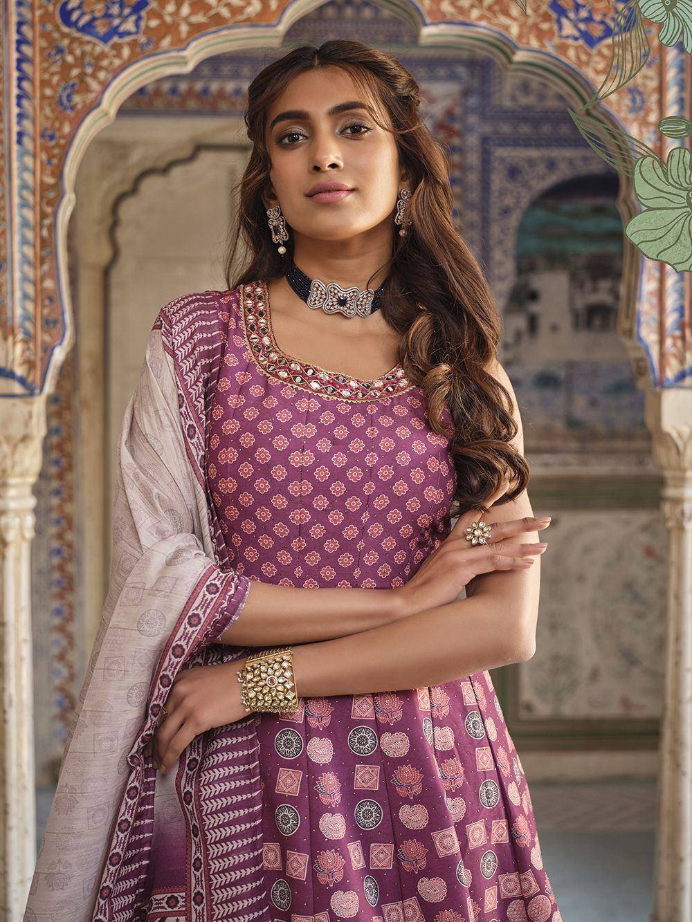Woman in purple floral print ethnic gown with dupatta, traditional jewelry, against ornate Indian architecture