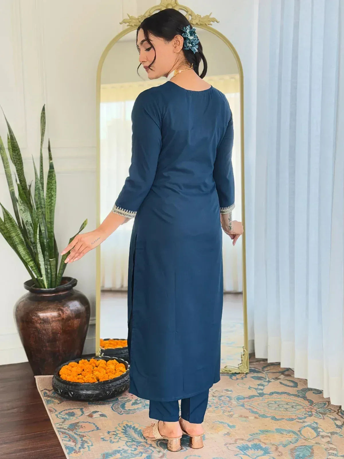 Woman in blue ethnic kurta with embroidered cuffs, paired with matching pants, standing near mirror and plant.