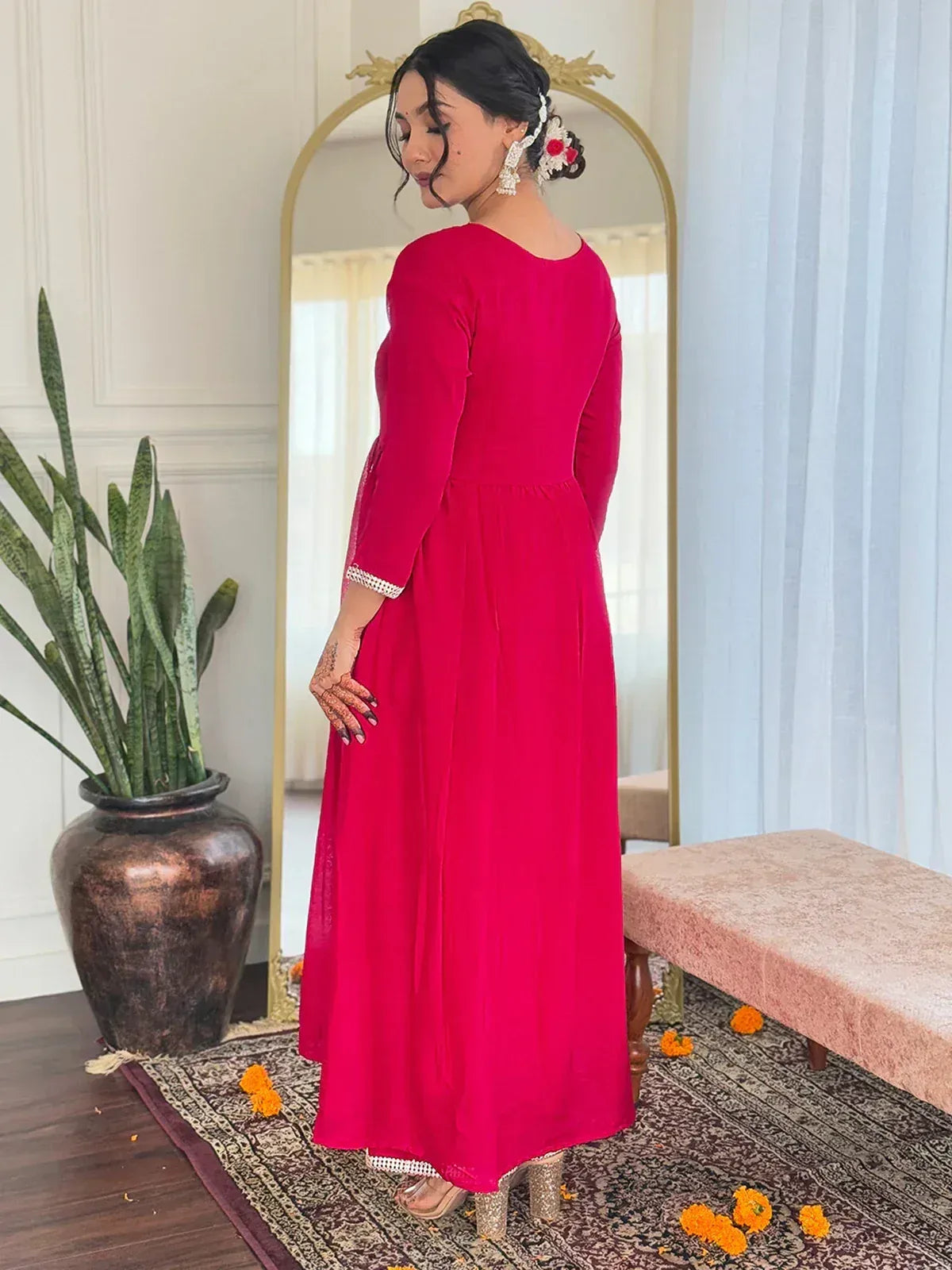 Woman in vibrant pink ethnic gown with lace cuffs, floral hair accessory, and henna hands, styled for Indian occasions.