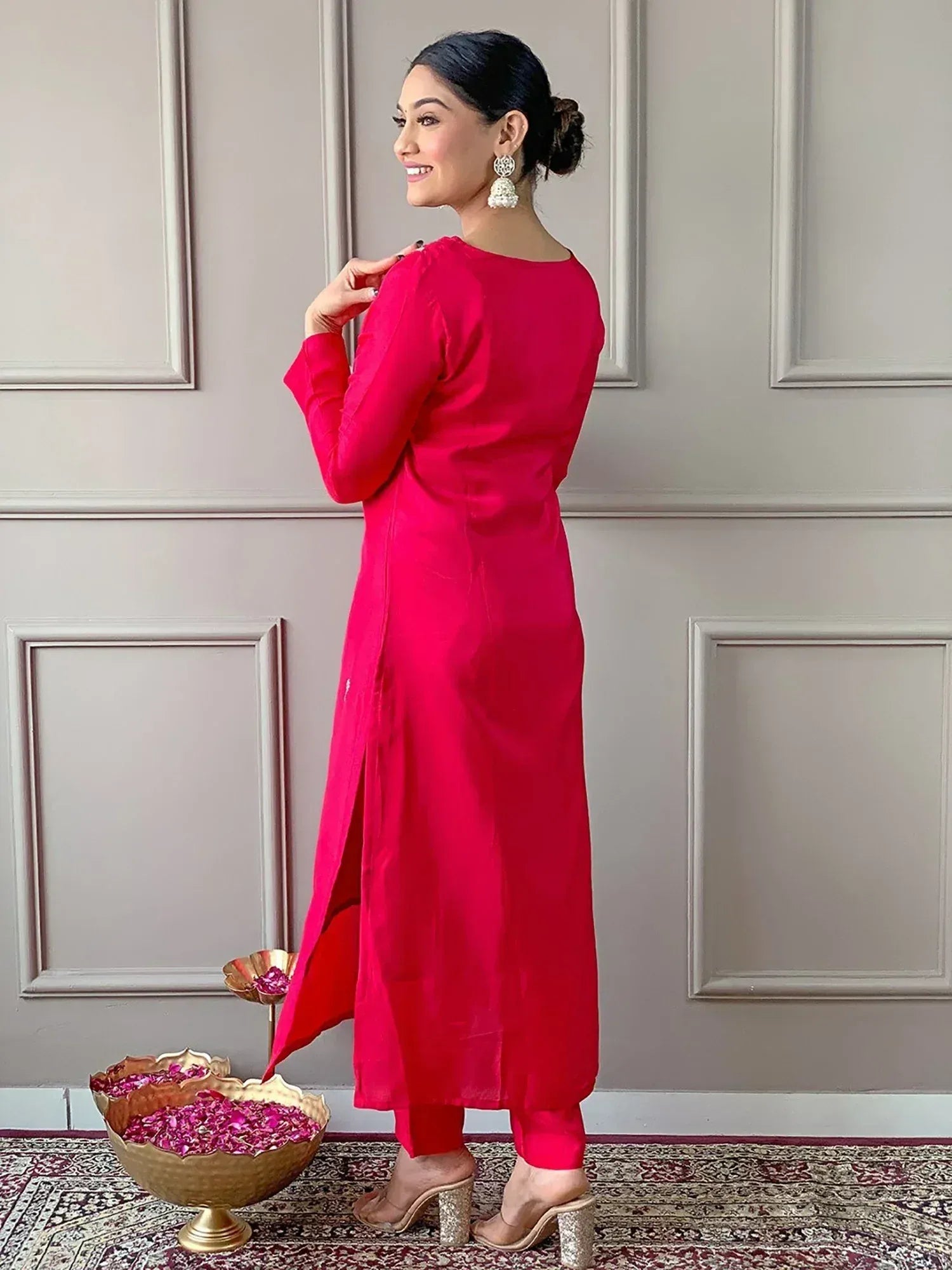 Woman in vibrant red ethnic kurta with side slit, paired with matching pants and jhumka earrings, standing near rose petals.