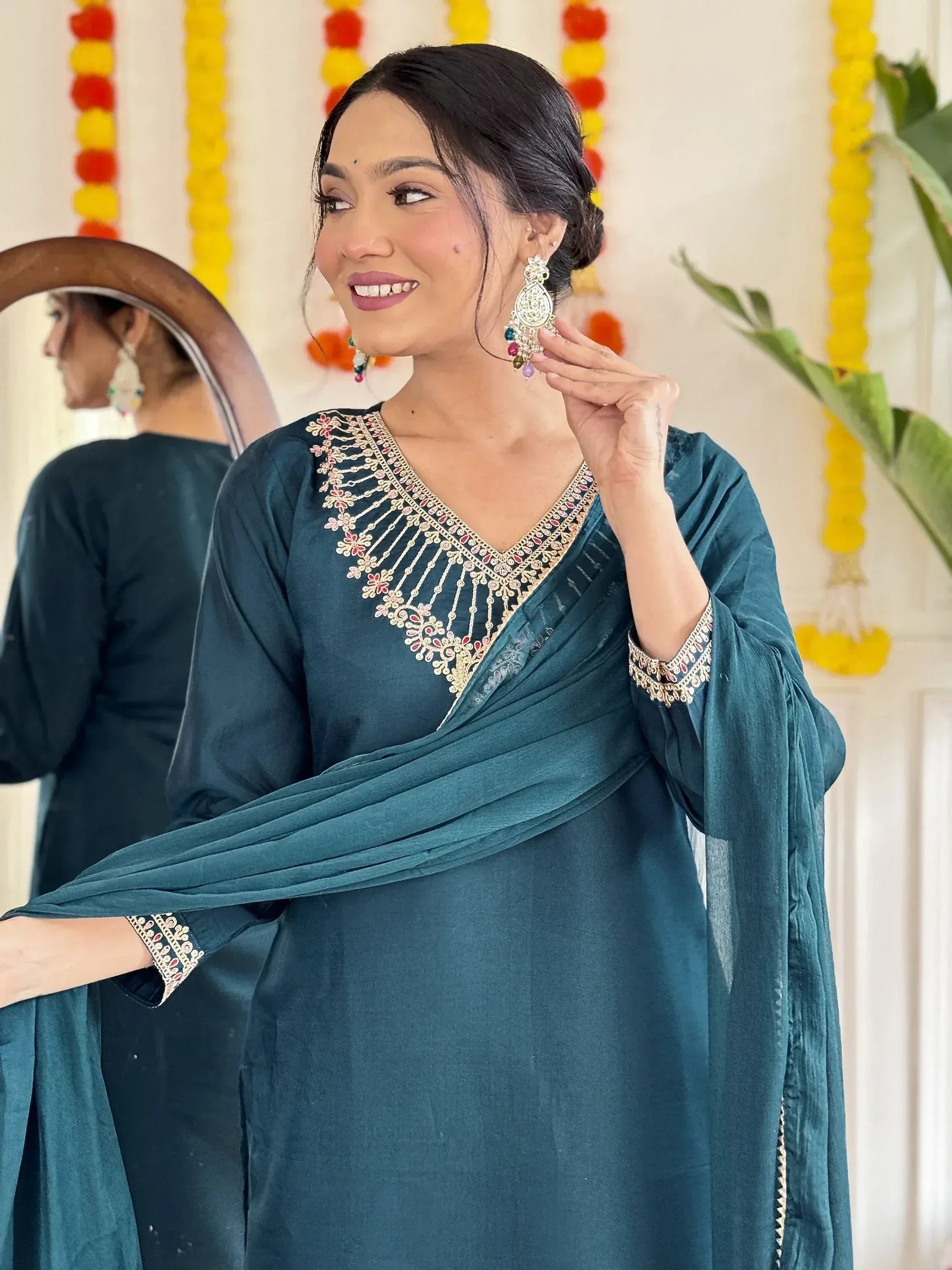 Woman in teal embroidered salwar kameez with mirror and floral decor, showcasing Indian ethnic wear.