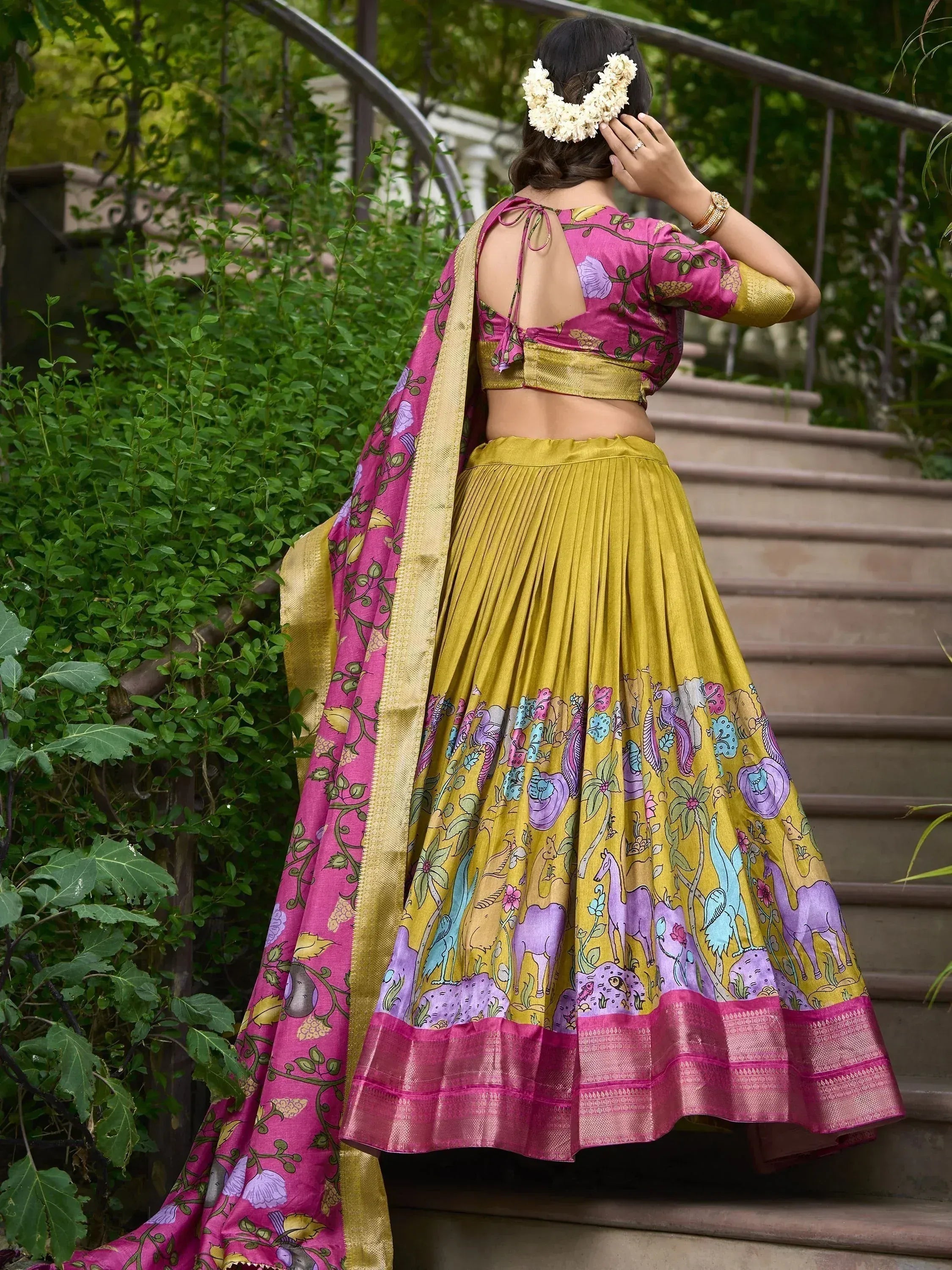 Woman in vibrant pink and gold printed lehenga choli with floral dupatta, standing on garden stairs