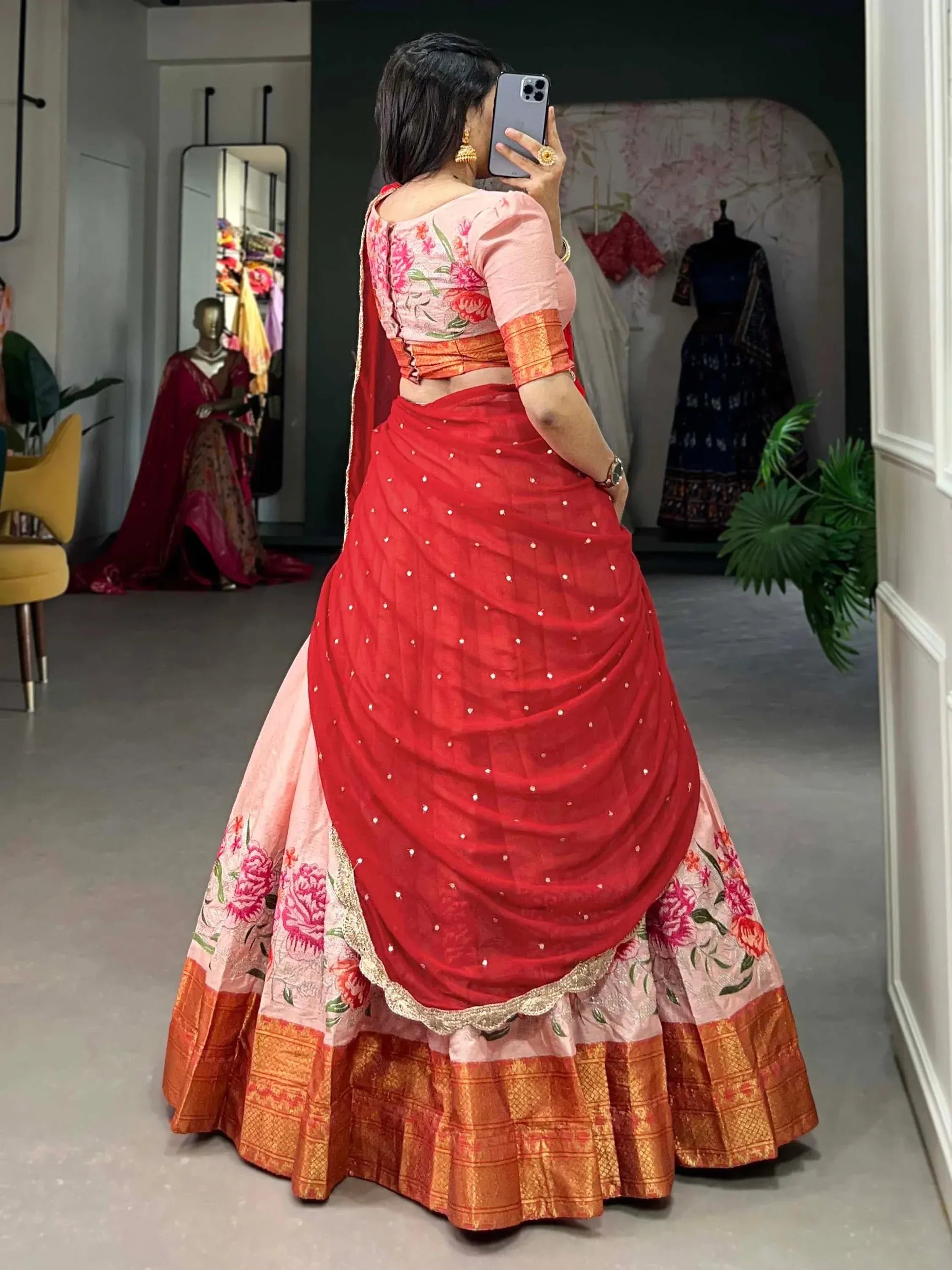 Woman in red and pink floral lehenga choli with gold border, taking mirror selfie in boutique