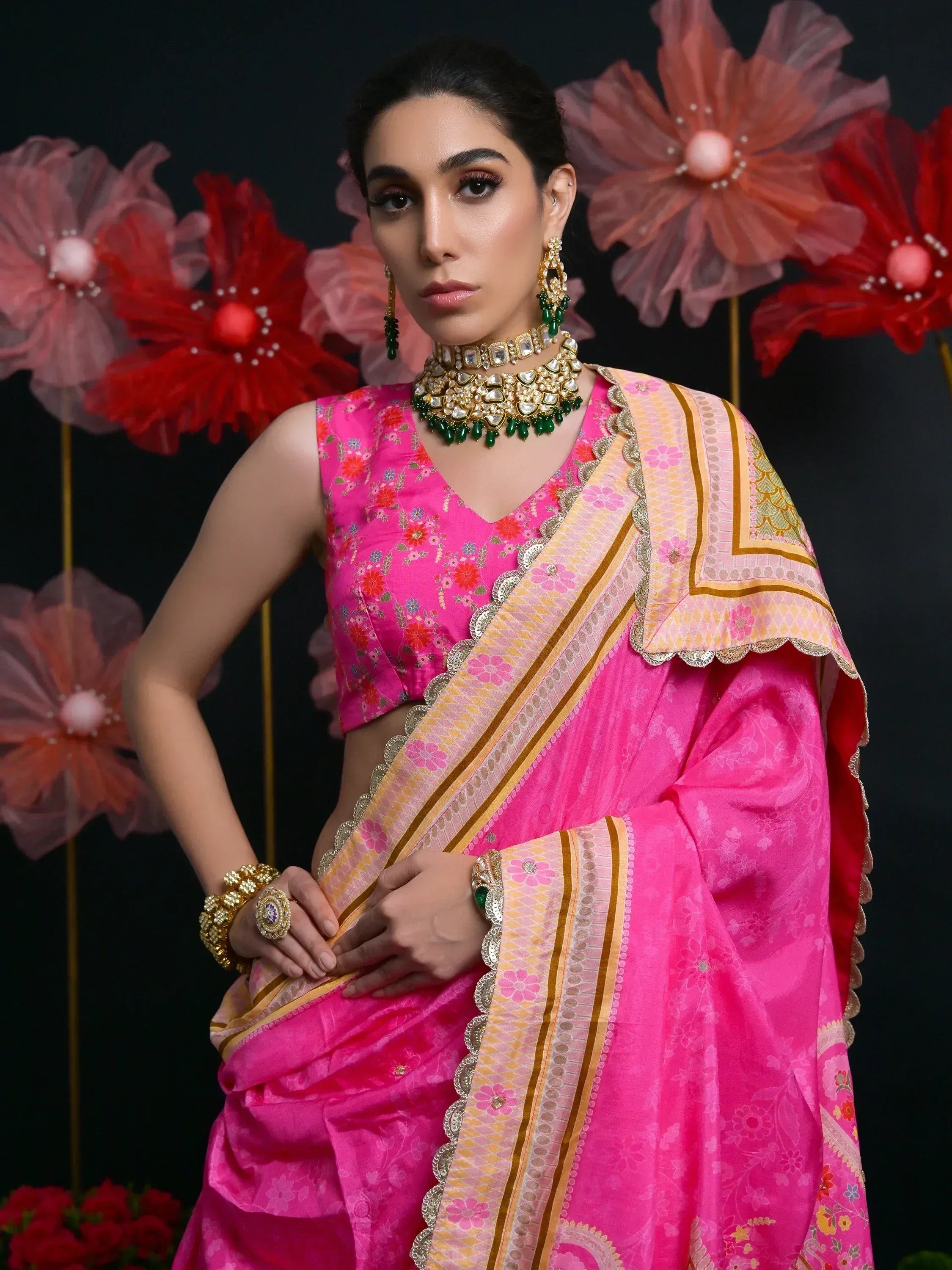 Woman in vibrant pink floral saree with gold border, paired with traditional Indian jewelry, perfect for festive occasions.