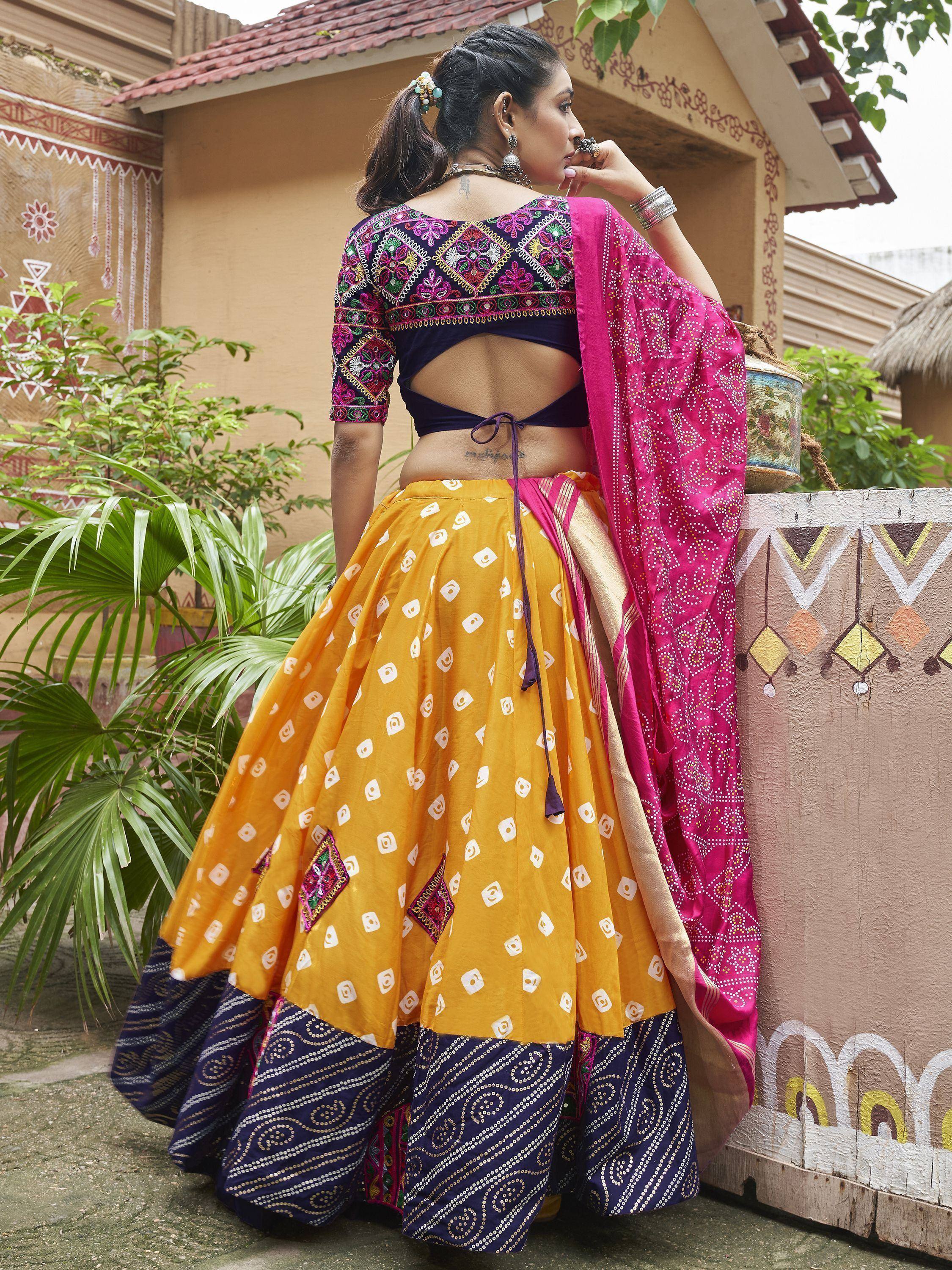 Woman in vibrant yellow and pink ethnic lehenga choli with mirror work, standing outdoors near painted wall