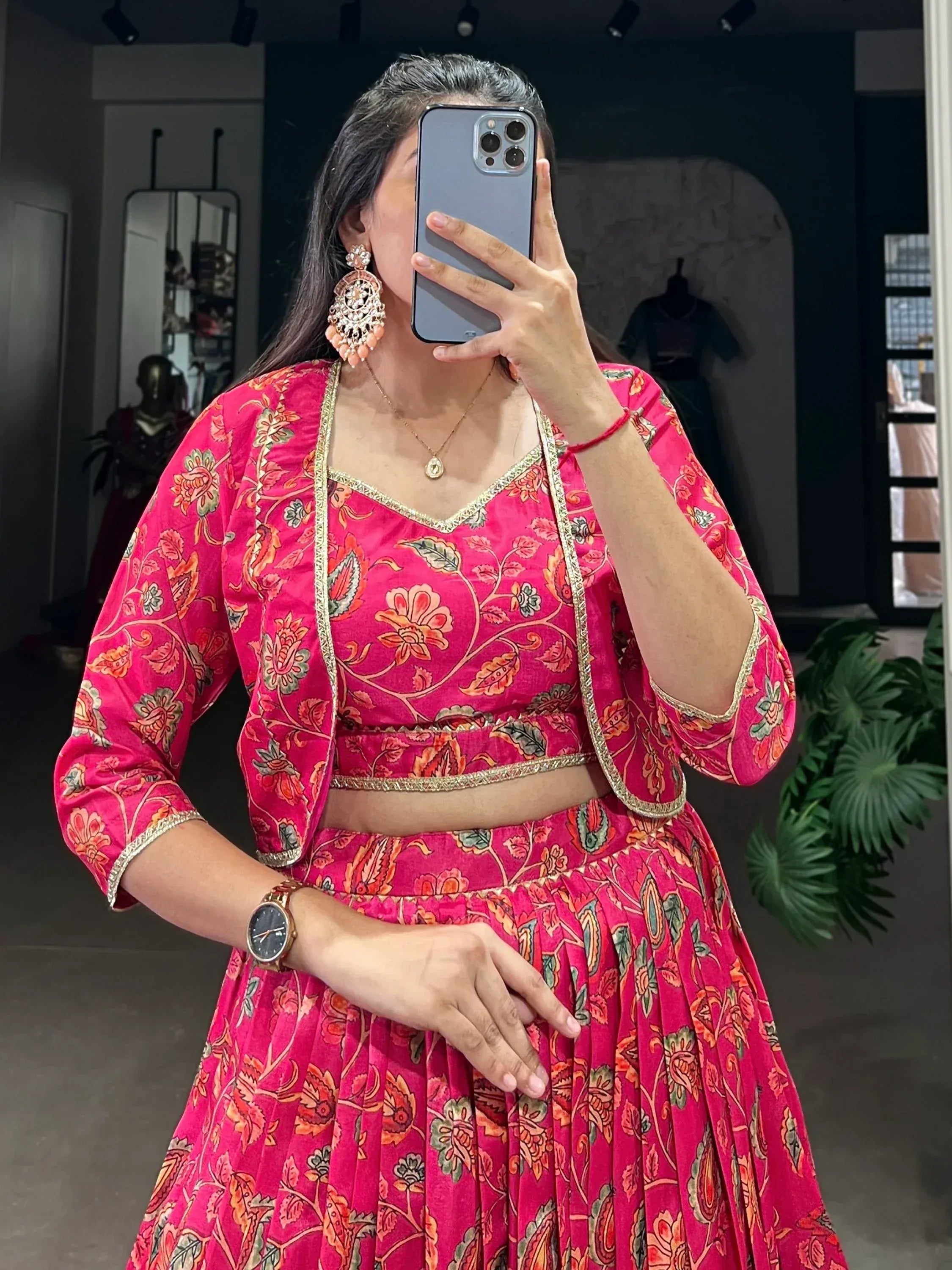 Woman in vibrant pink floral lehenga choli with gold trim, taking mirror selfie in boutique