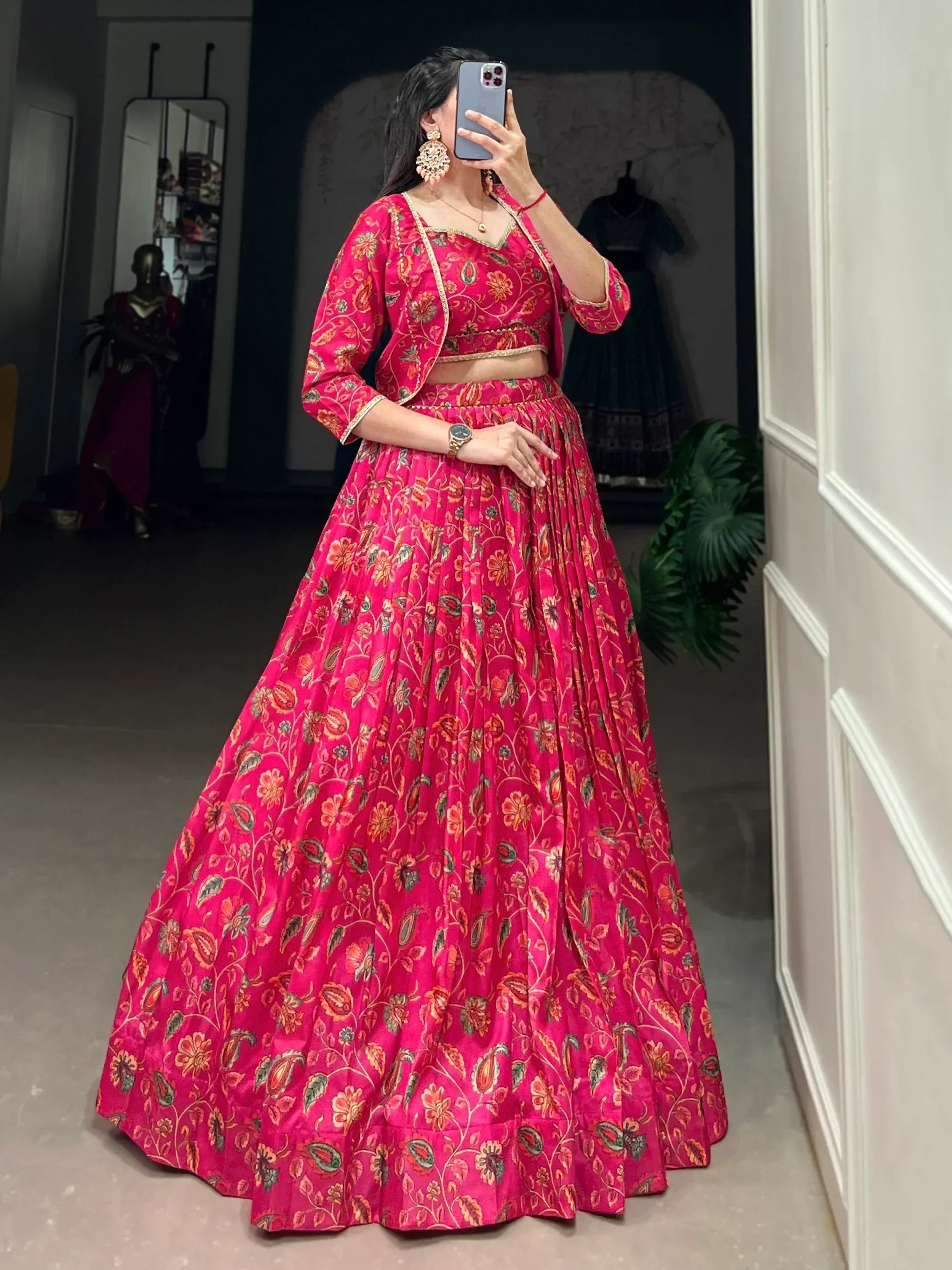Woman in vibrant pink floral lehenga choli with gold trim, taking mirror selfie in boutique