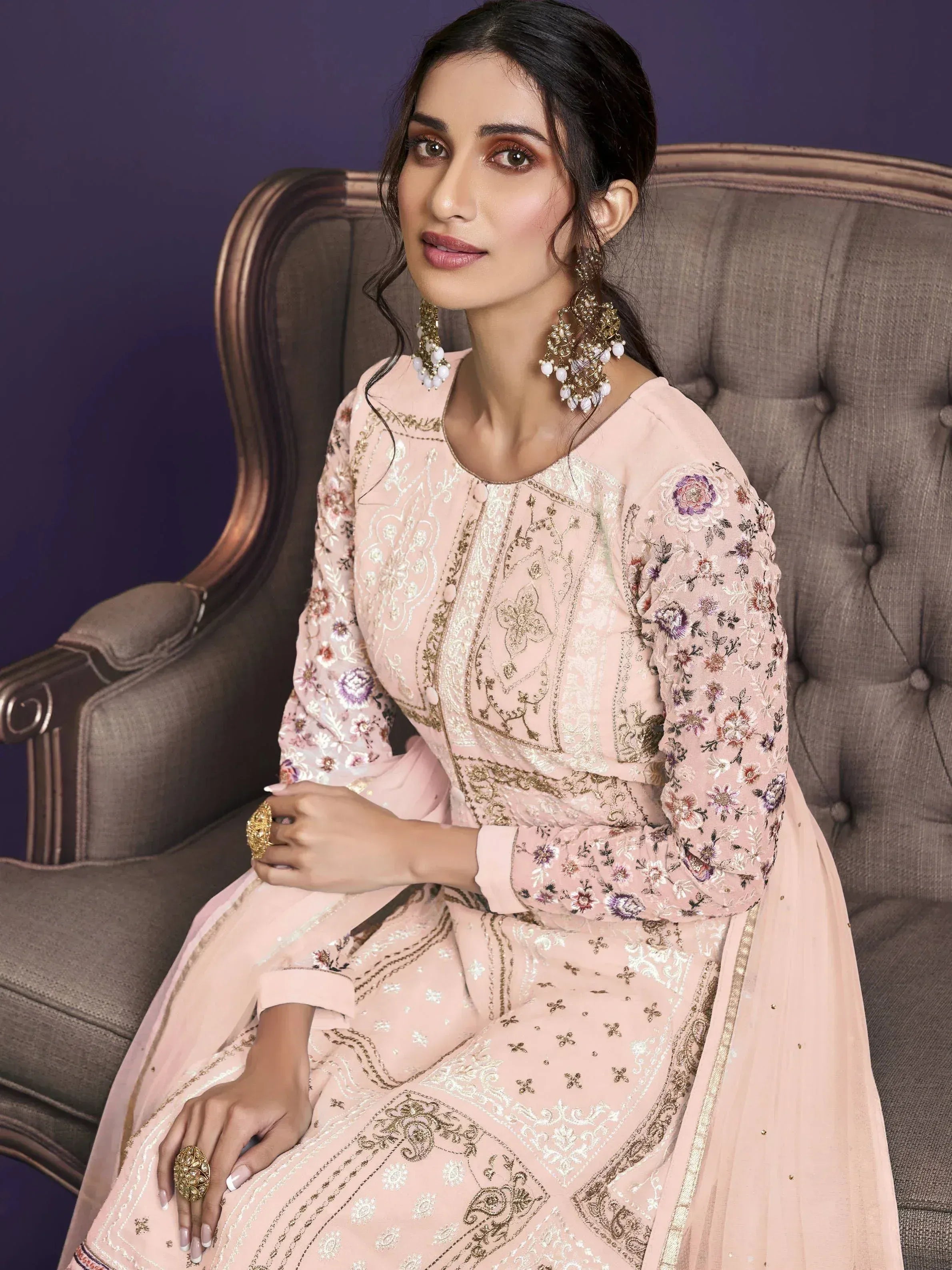 Woman in elegant pastel pink embroidered salwar kameez with statement earrings, seated on a classic chair