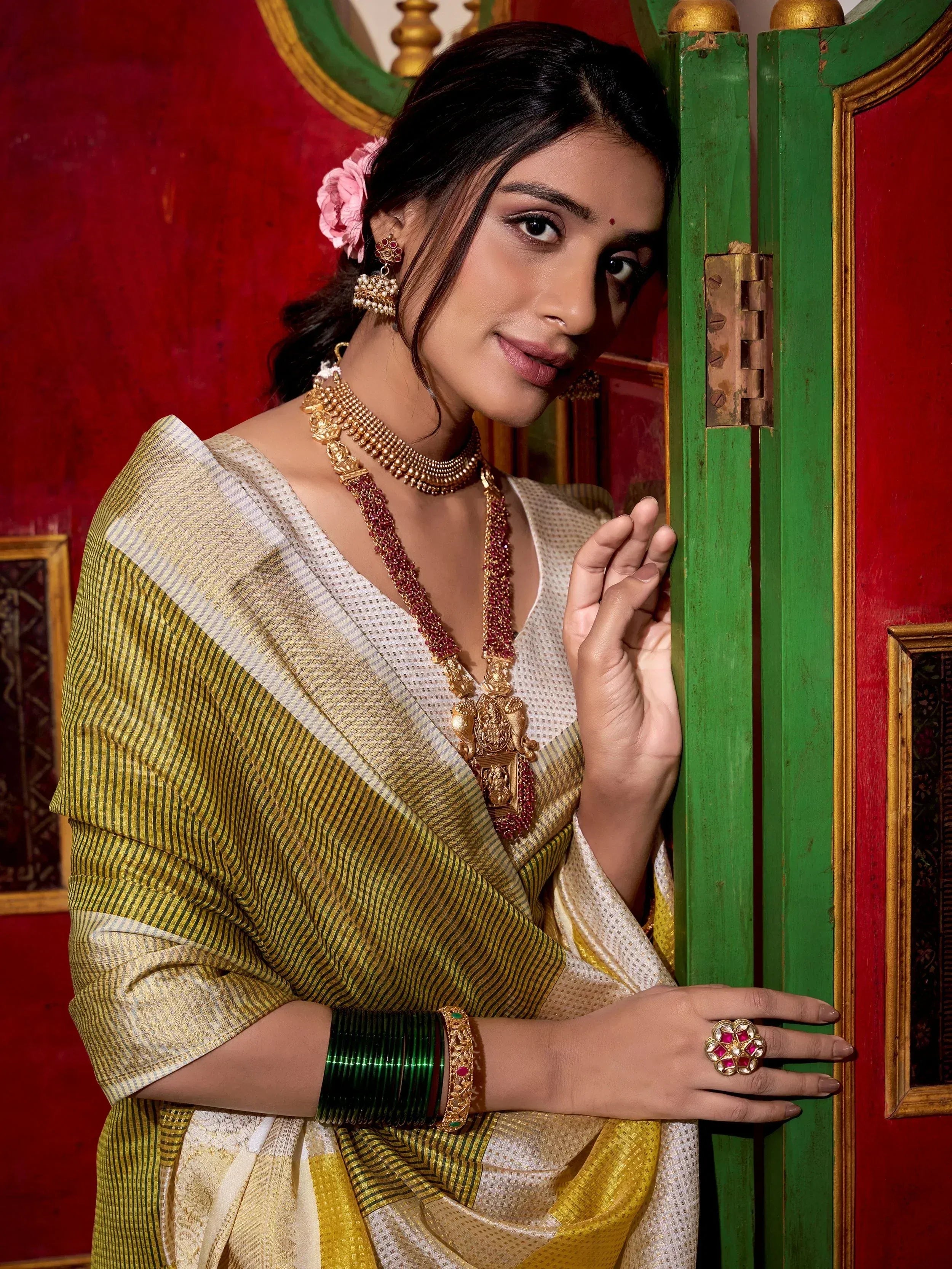 Woman in traditional Indian saree with gold jewelry, green bangles, and floral hair accessory against red and green backdrop