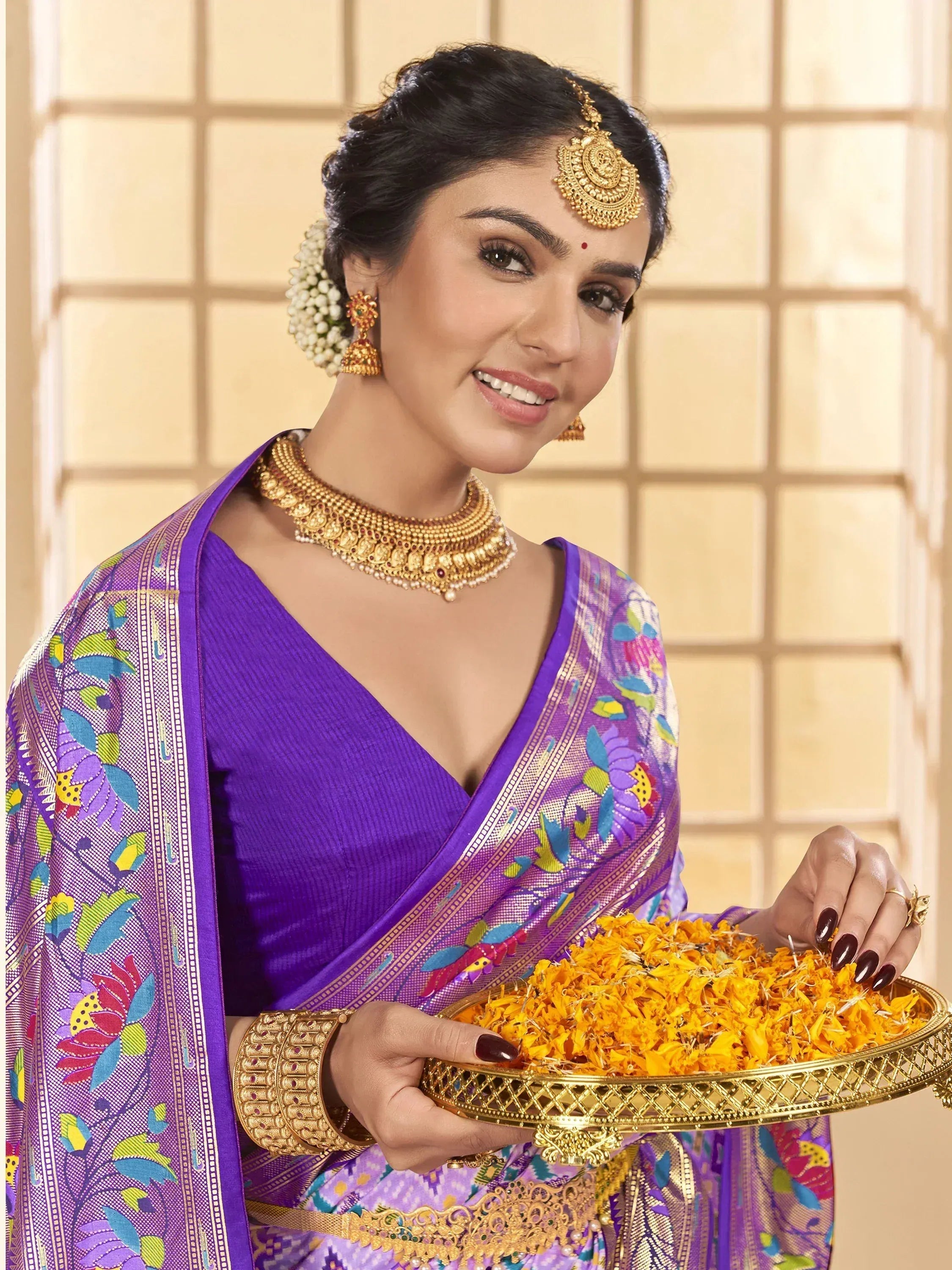 Woman in purple floral saree with gold jewelry holding marigold flowers, traditional Indian ethnic wear