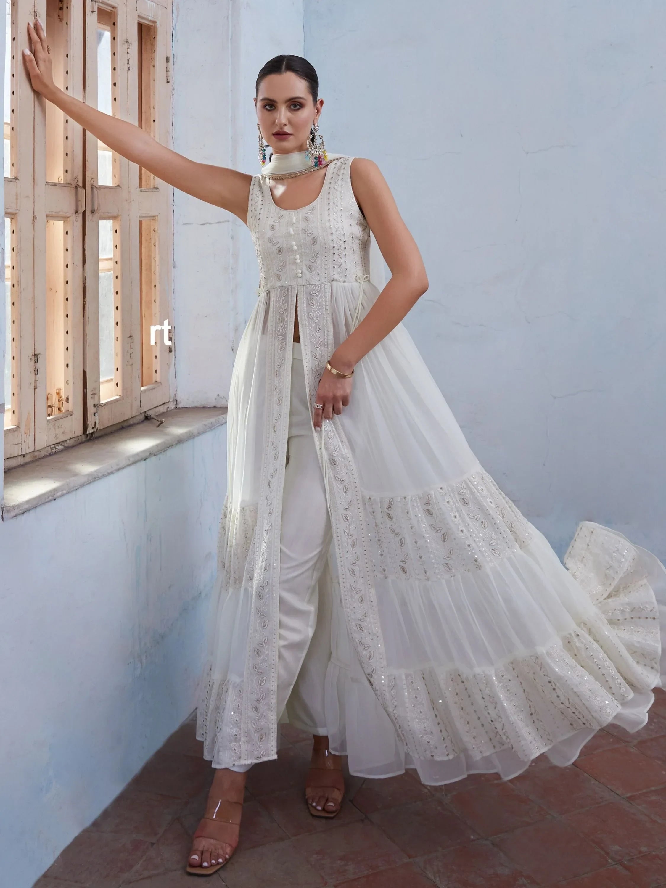 Woman in white embroidered ethnic gown with sheer layers, standing by a window
