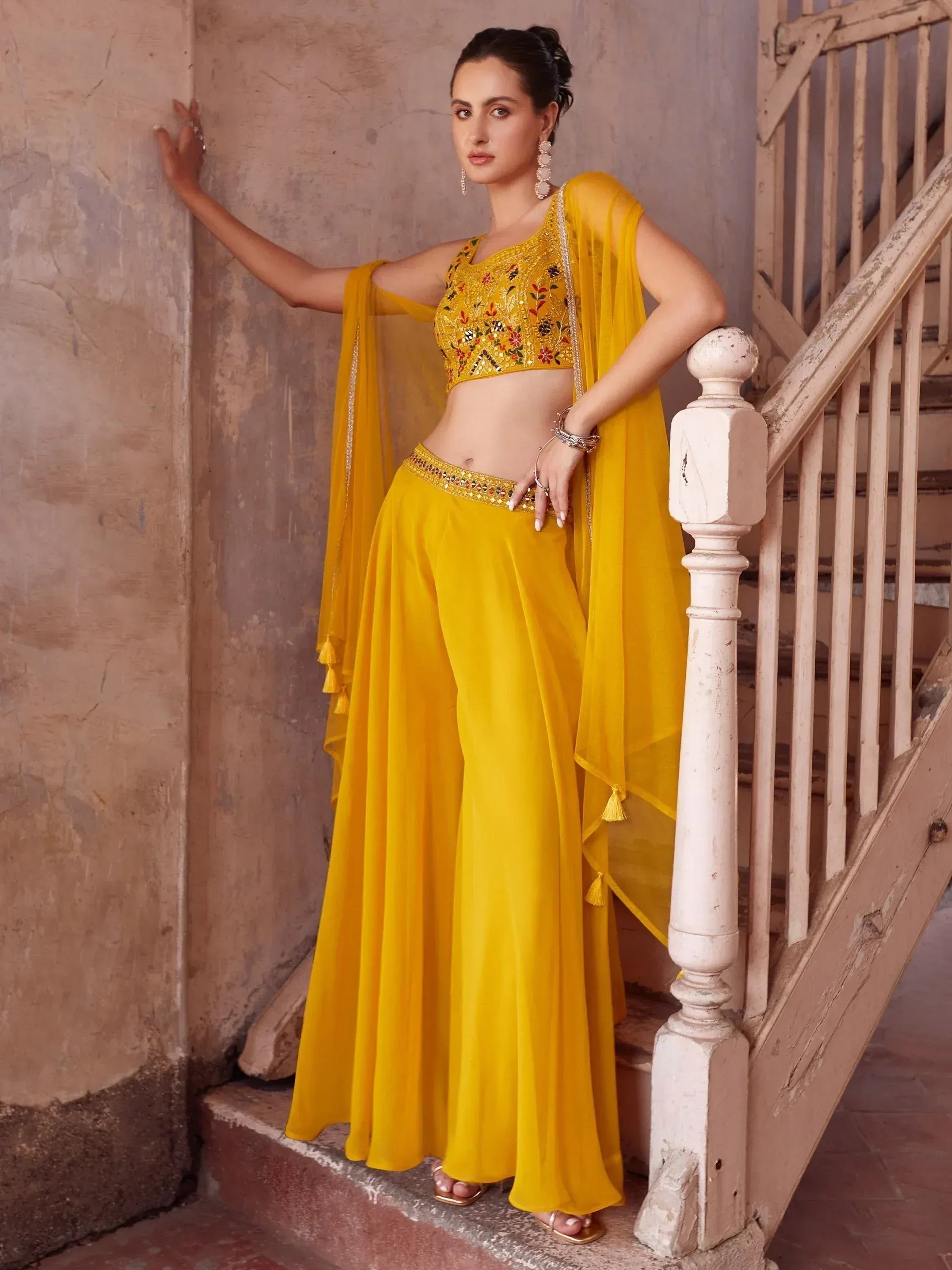 Woman wearing yellow embroidered lehenga choli set with dupatta, posing by staircase