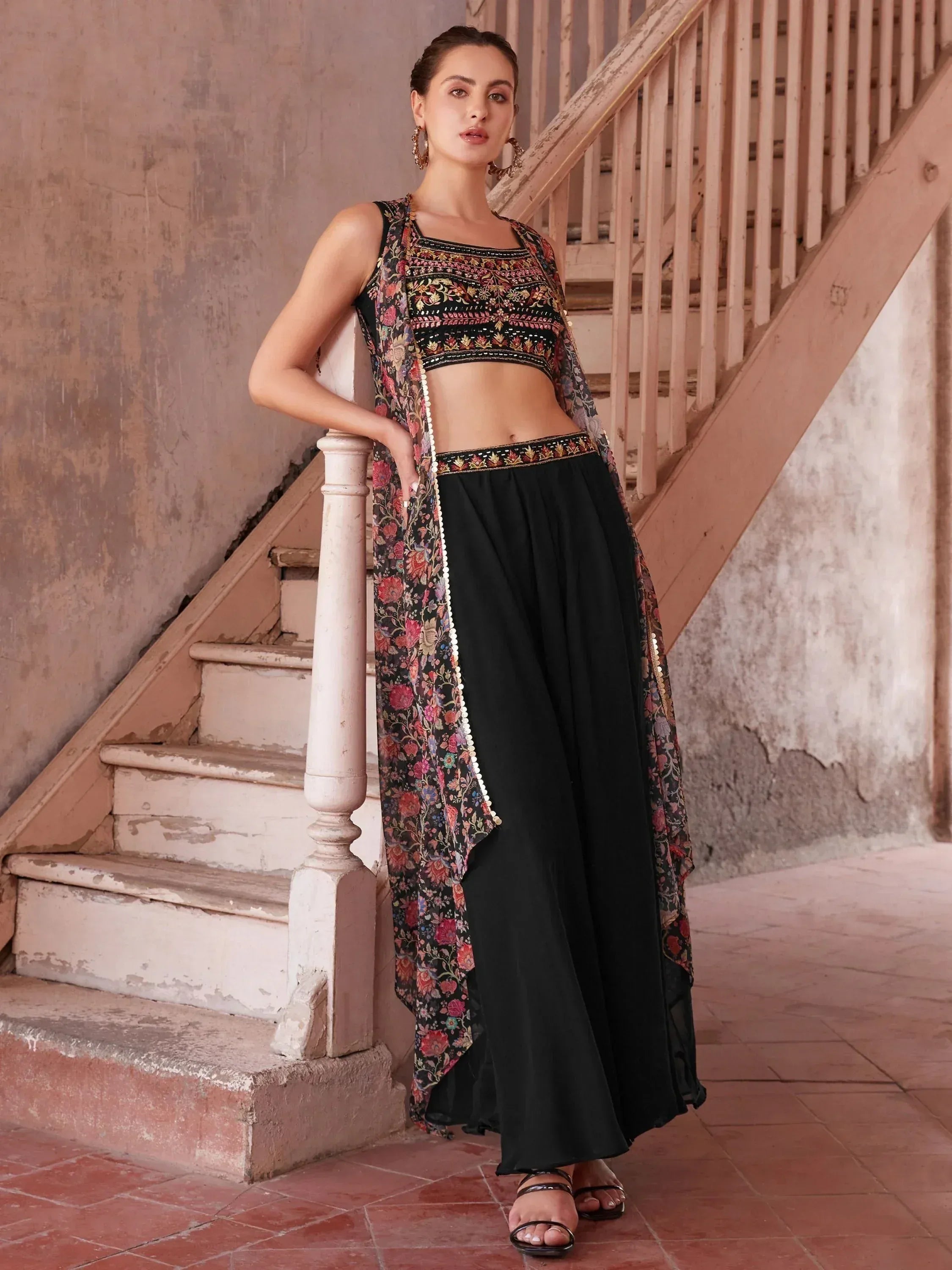 Woman wearing black floral embroidered ethnic crop top and palazzo pants set with a matching long shrug, standing on rustic stairs.